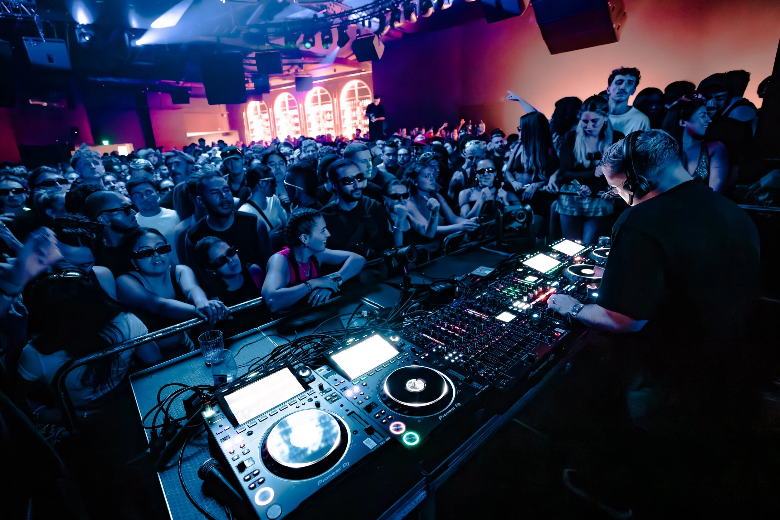 Crowd at a nightclub with a DJ playing electronic music, colorful neon lights, and people dancing and enjoying the event.