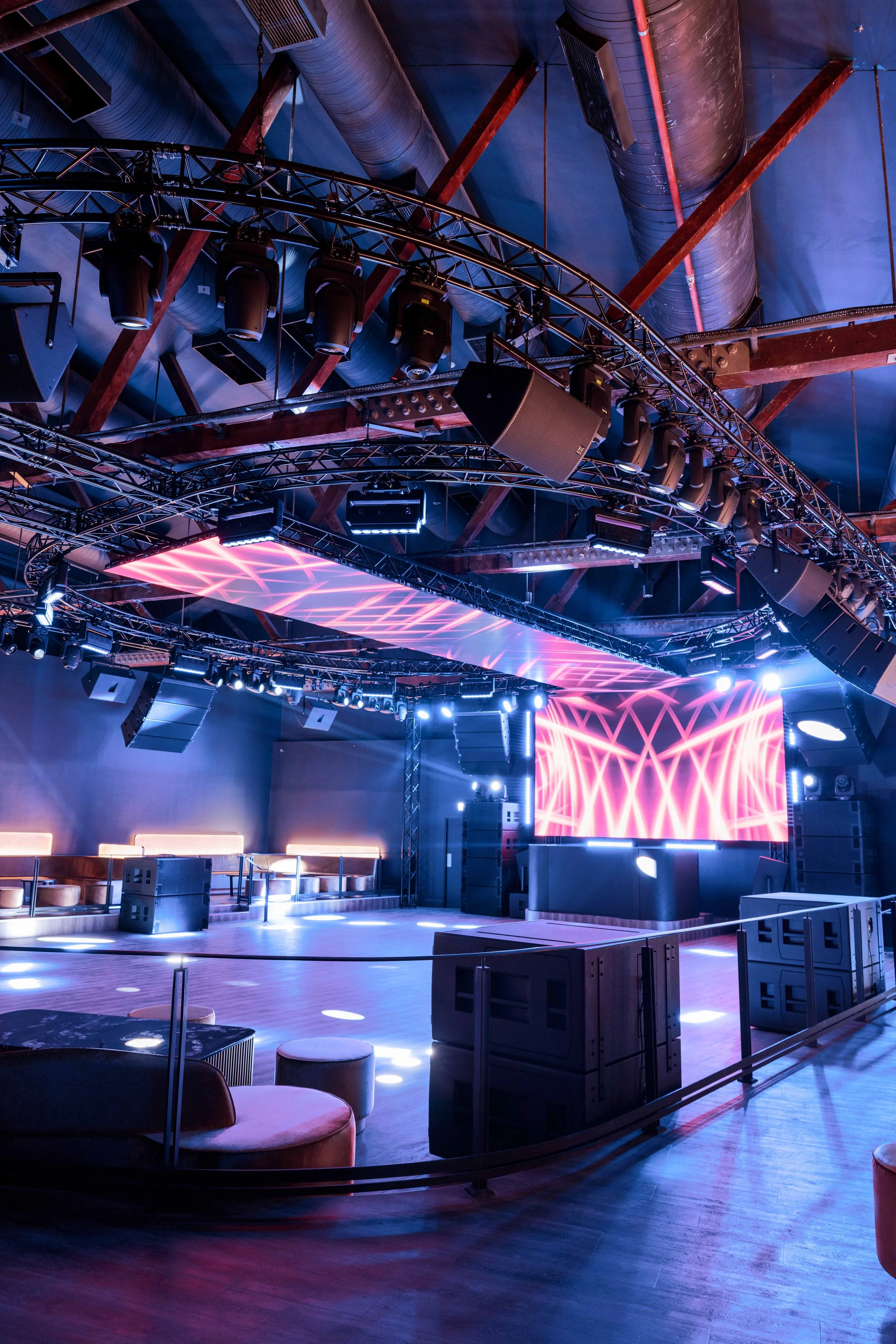 Empty nightclub with a large LED screen displaying pink geometric patterns, surrounded by stage lighting and audio equipment.
