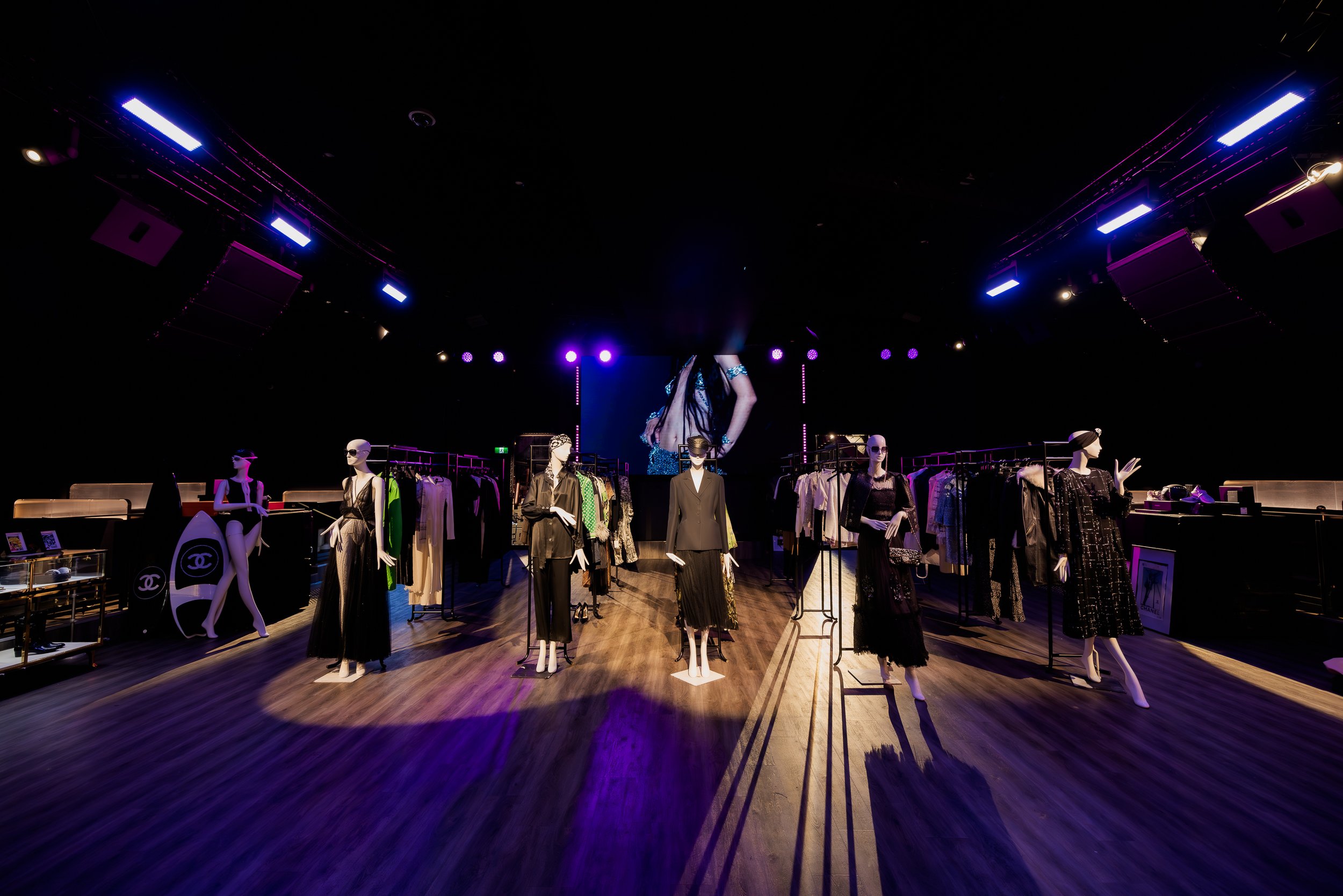 Mannequin display with stylish black and beige outfits, sunglasses, and handbags, set in a boutique with clothing racks and a large digital screen in the background.