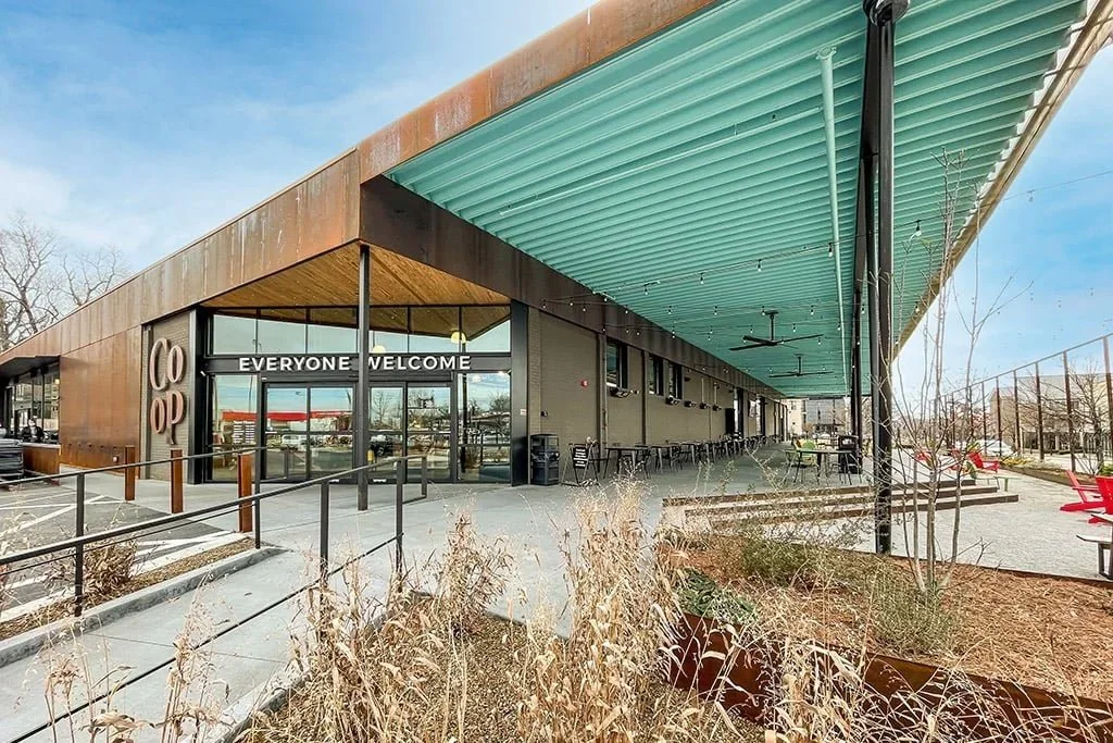 Modern commercial building with a rust-colored exterior and teal roof, featuring glass doors with a sign that says "Everyone Welcome," outdoor seating area with black chairs and tables, and a landscaped yard with dry plants.