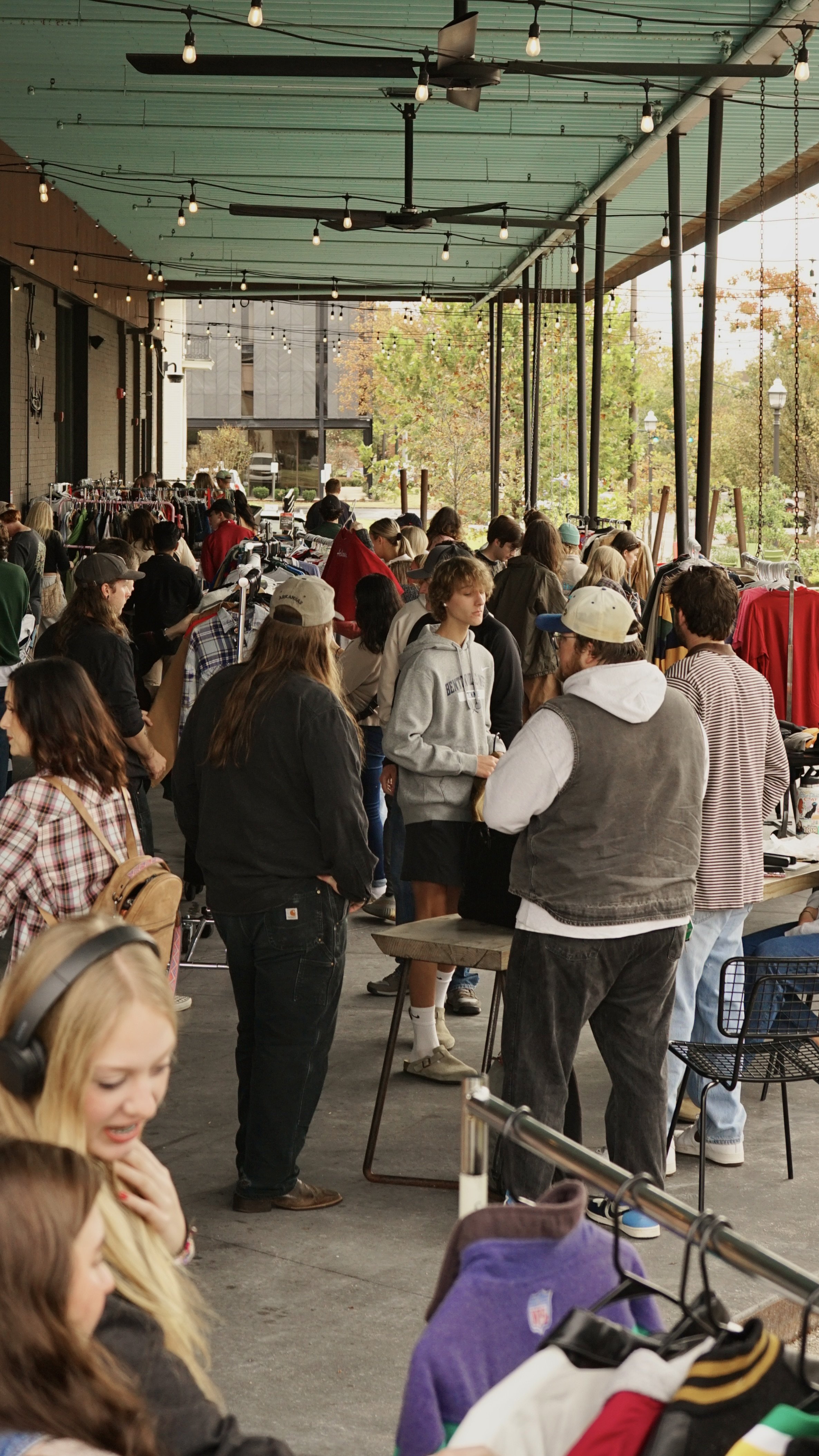 People browsing clothes at an outdoor market under string lights, with trees and buildings visible in the background.