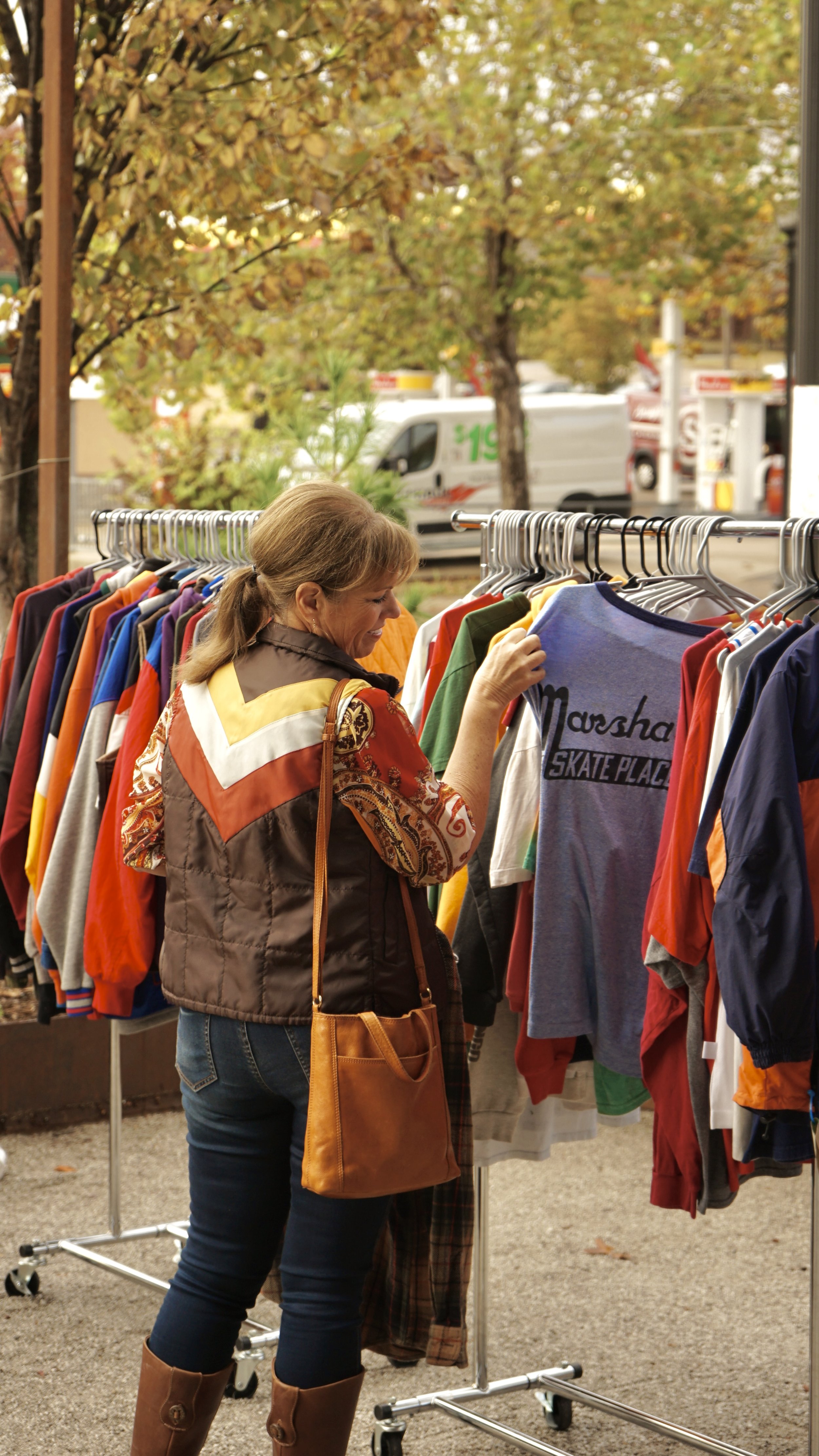Woman shopping outdoors, browsing through a rack of colorful clothes, with trees and a street scene with trucks in the background.