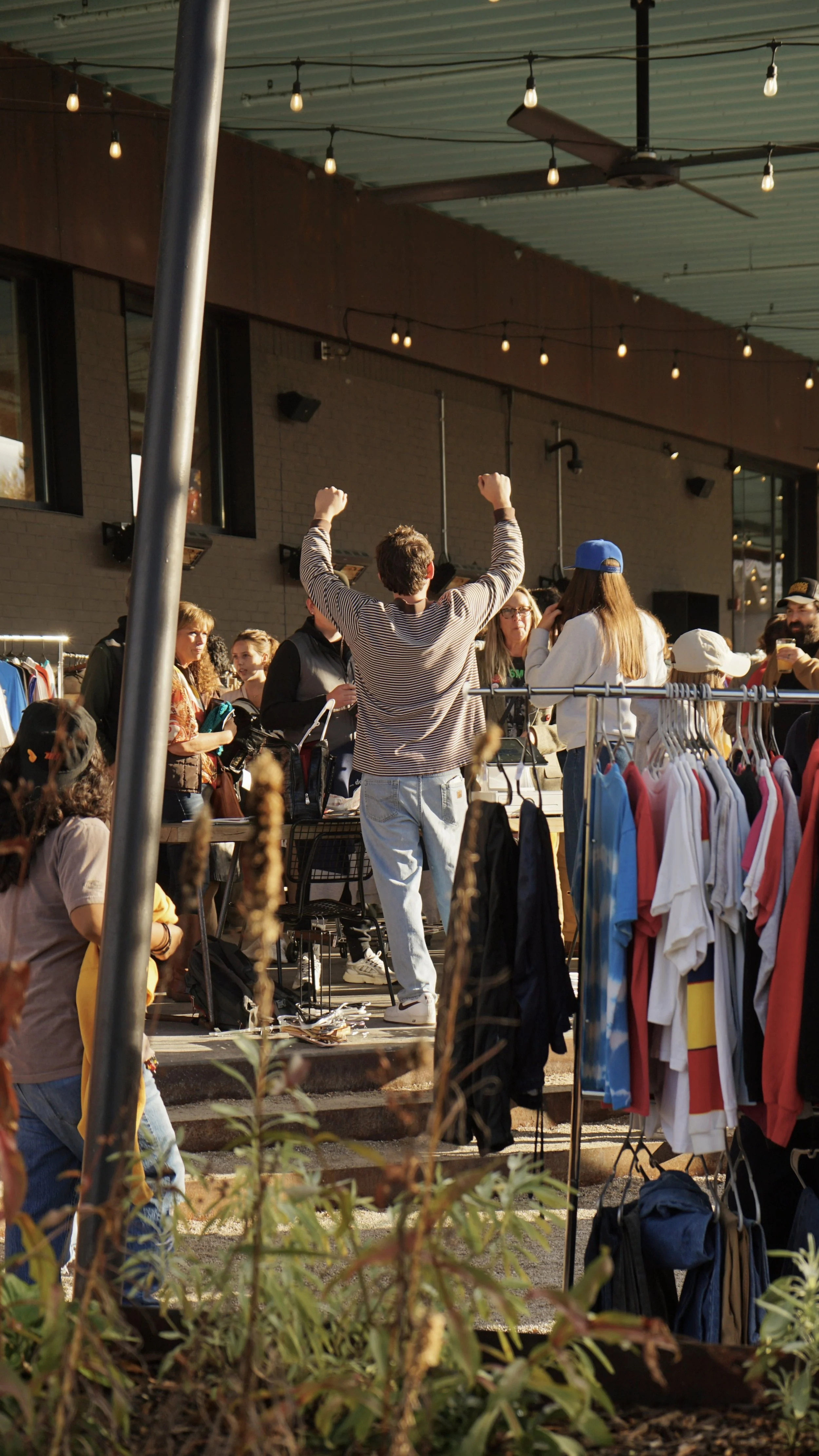 A group of people at an outdoor market with clothing and accessories on display. A man with curly hair, wearing a striped shirt and gray jeans, is standing with arms raised. The market is decorated with string lights and located under a building with large windows and a corrugated metal roof.