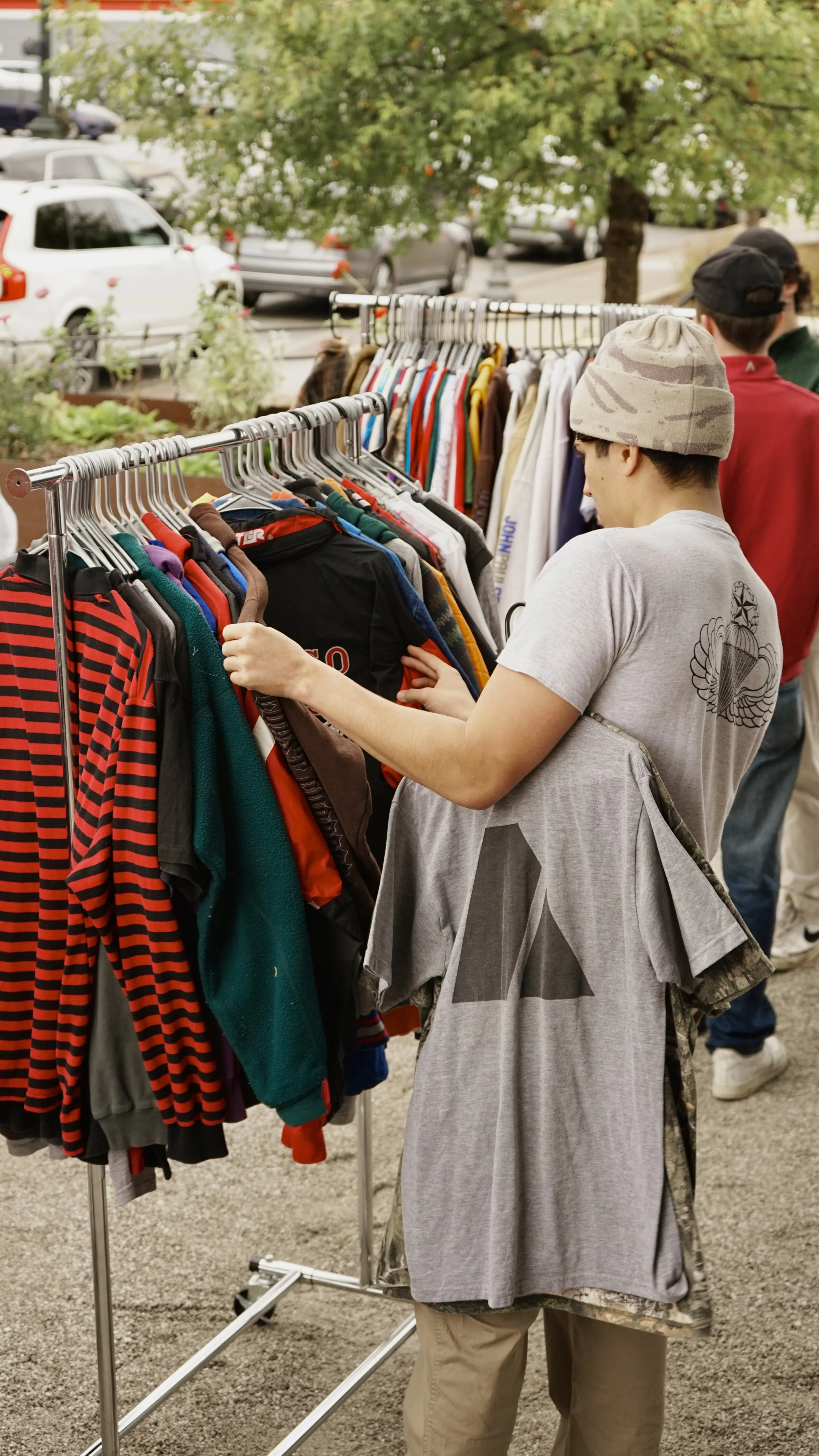 People browsing through clothing on racks at an outdoor market with cars and trees in the background.