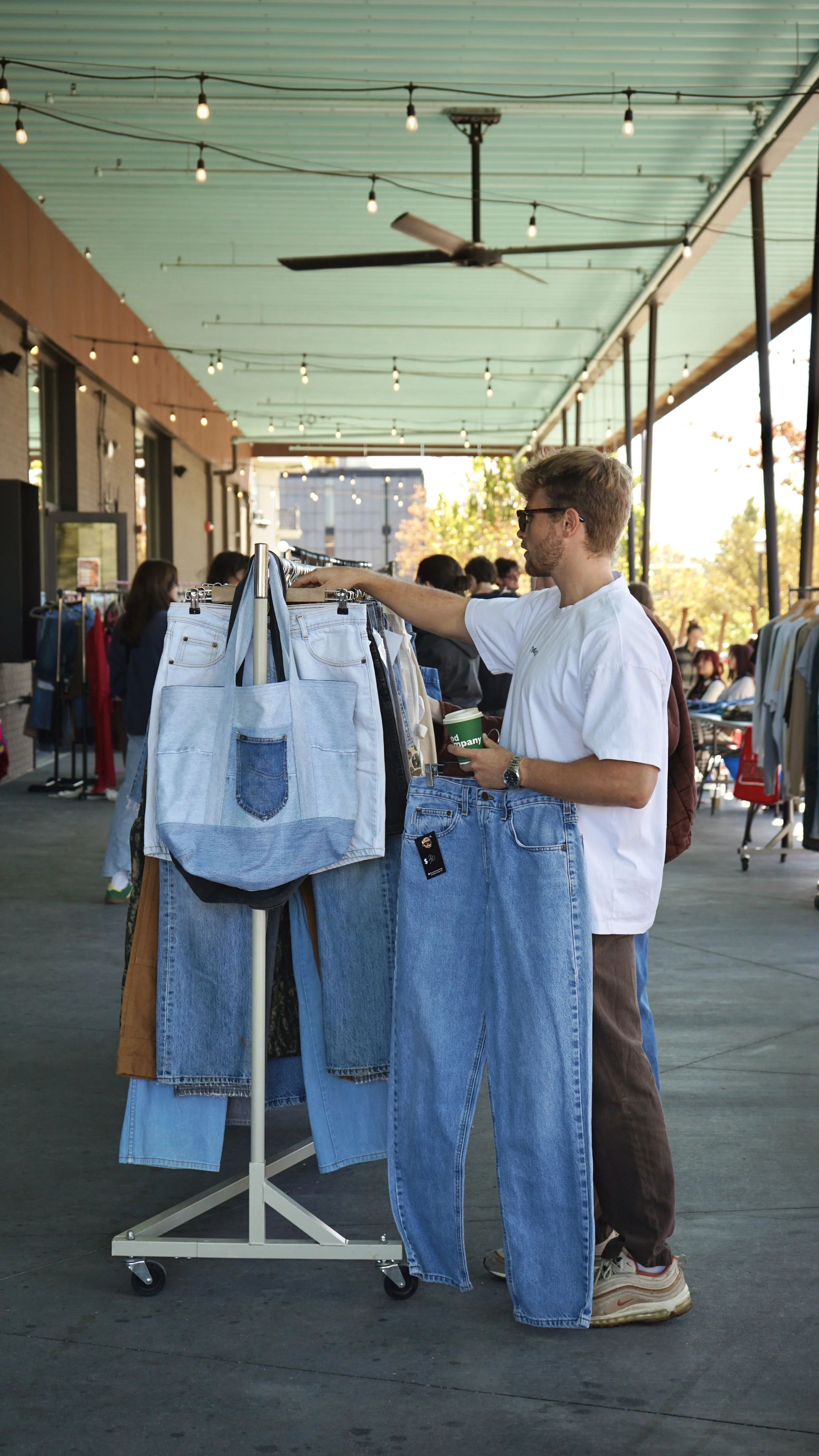 A man in a white t-shirt and jeans shopping at an outdoor market, browsing a clothing rack filled with denim jeans and bags.