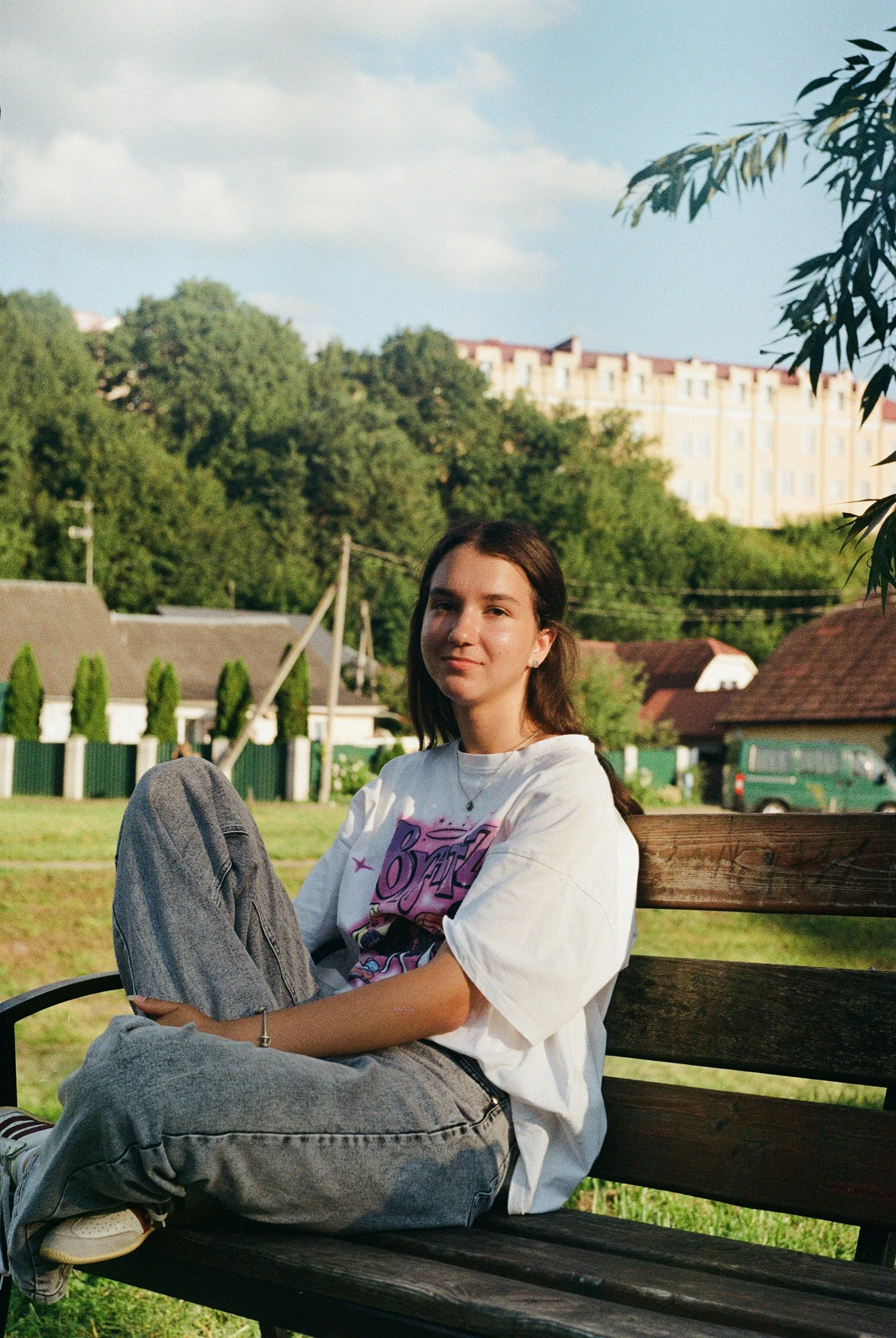 Young woman sitting on a park bench outdoors with trees and buildings in the background.