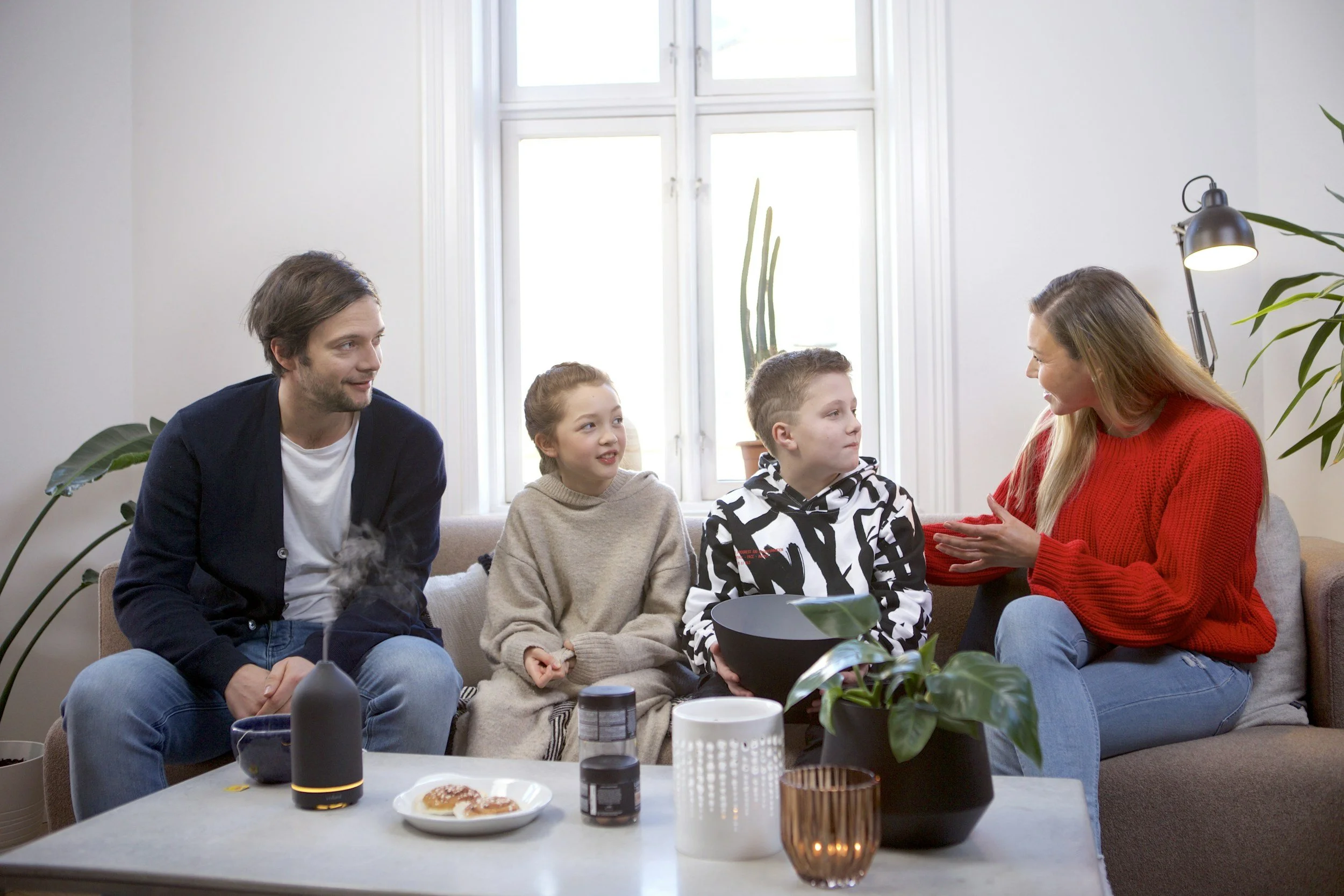 Family of four sitting together on a couch and talking in a bright living room.