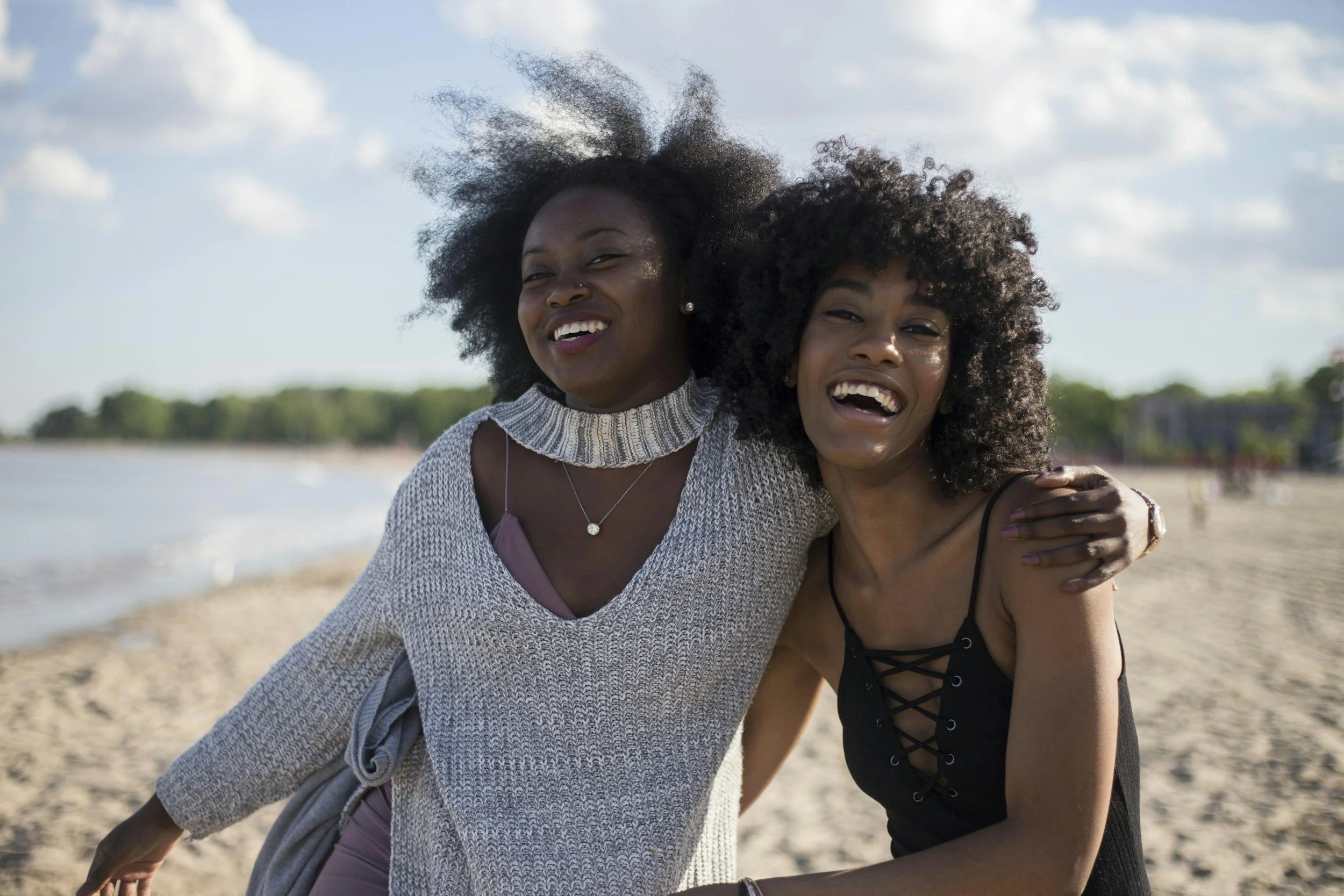 Two women smiling with their arms around each other while standing near the water.