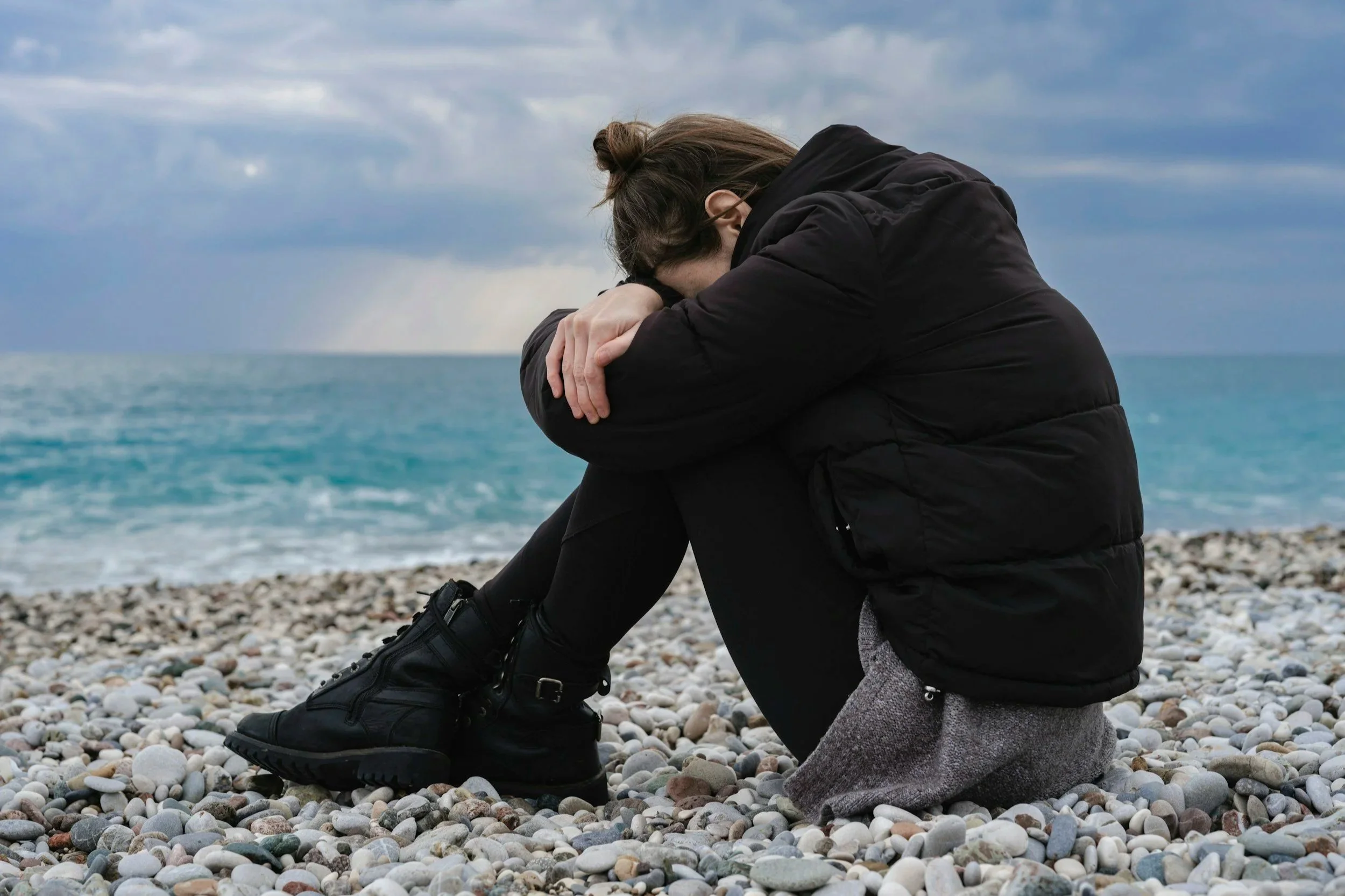A person sitting alone on a rocky beach with their head resting on their arms under a cloudy sky.