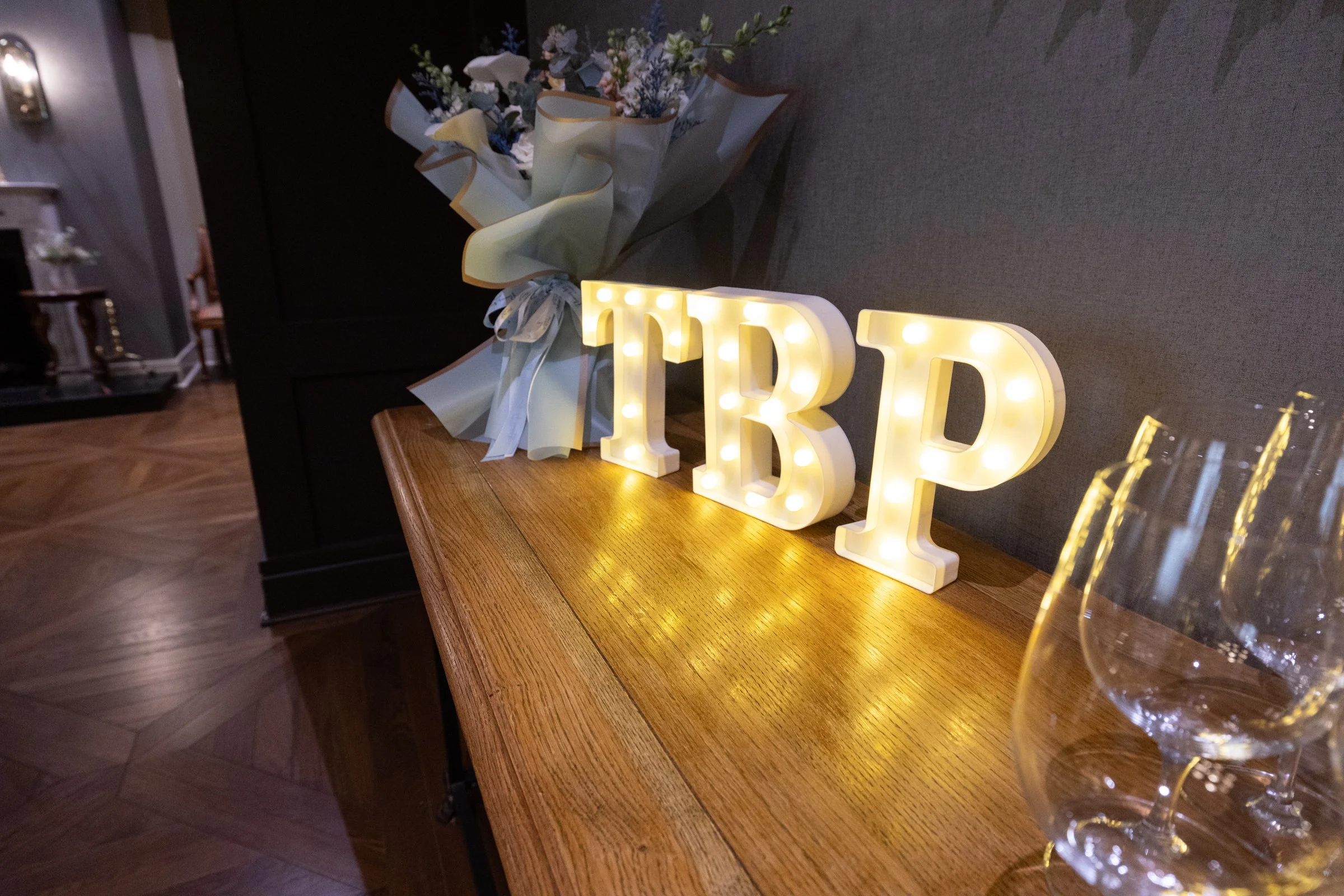 Decorative illuminated letters spelling 'RDP', a floral bouquet wrapped in paper, and a glass of champagne on a wooden table at a celebration event.