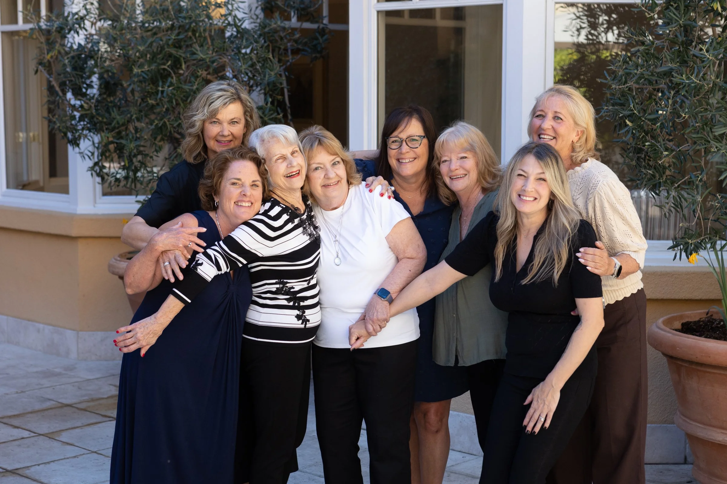 A group of nine women, mostly middle-aged to elderly, smiling and embracing each other outside near a house with large windows and potted plants.