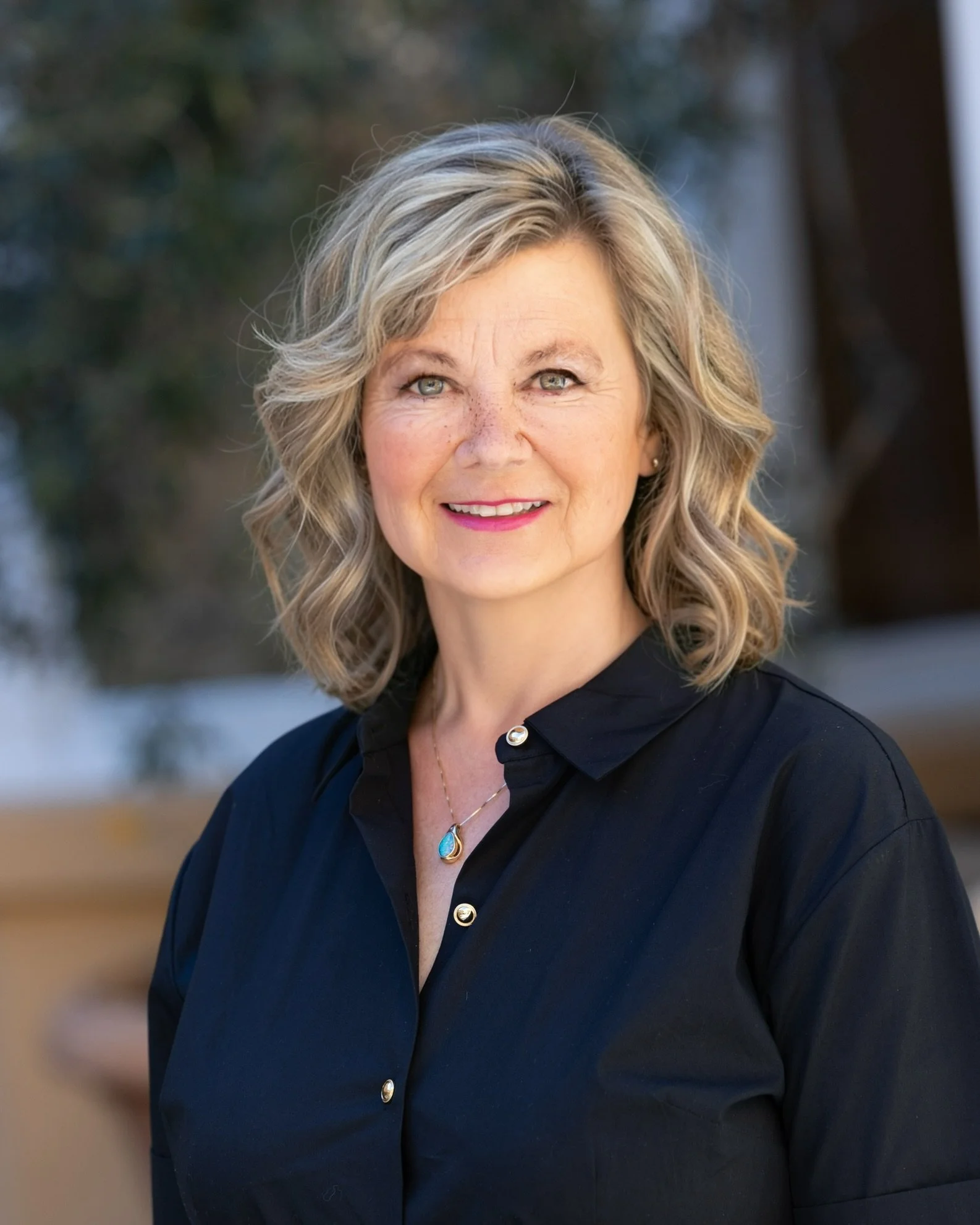 Portrait of a middle-aged woman with curly blond hair, wearing a black blouse with gold buttons, and a necklace with a blue pendant.
