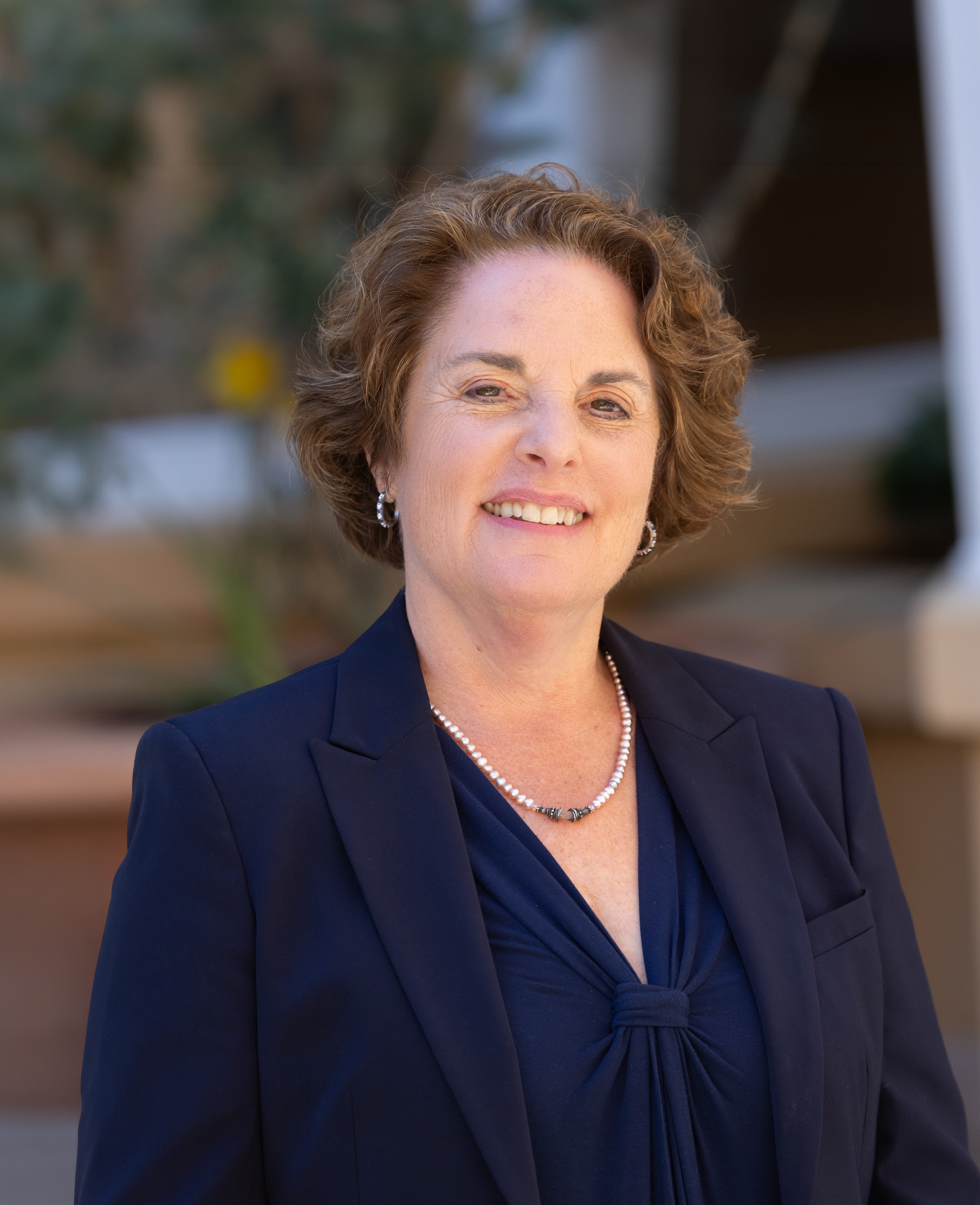 A woman with short curly hair smiling and wearing a navy blue blazer and a pearl necklace.
