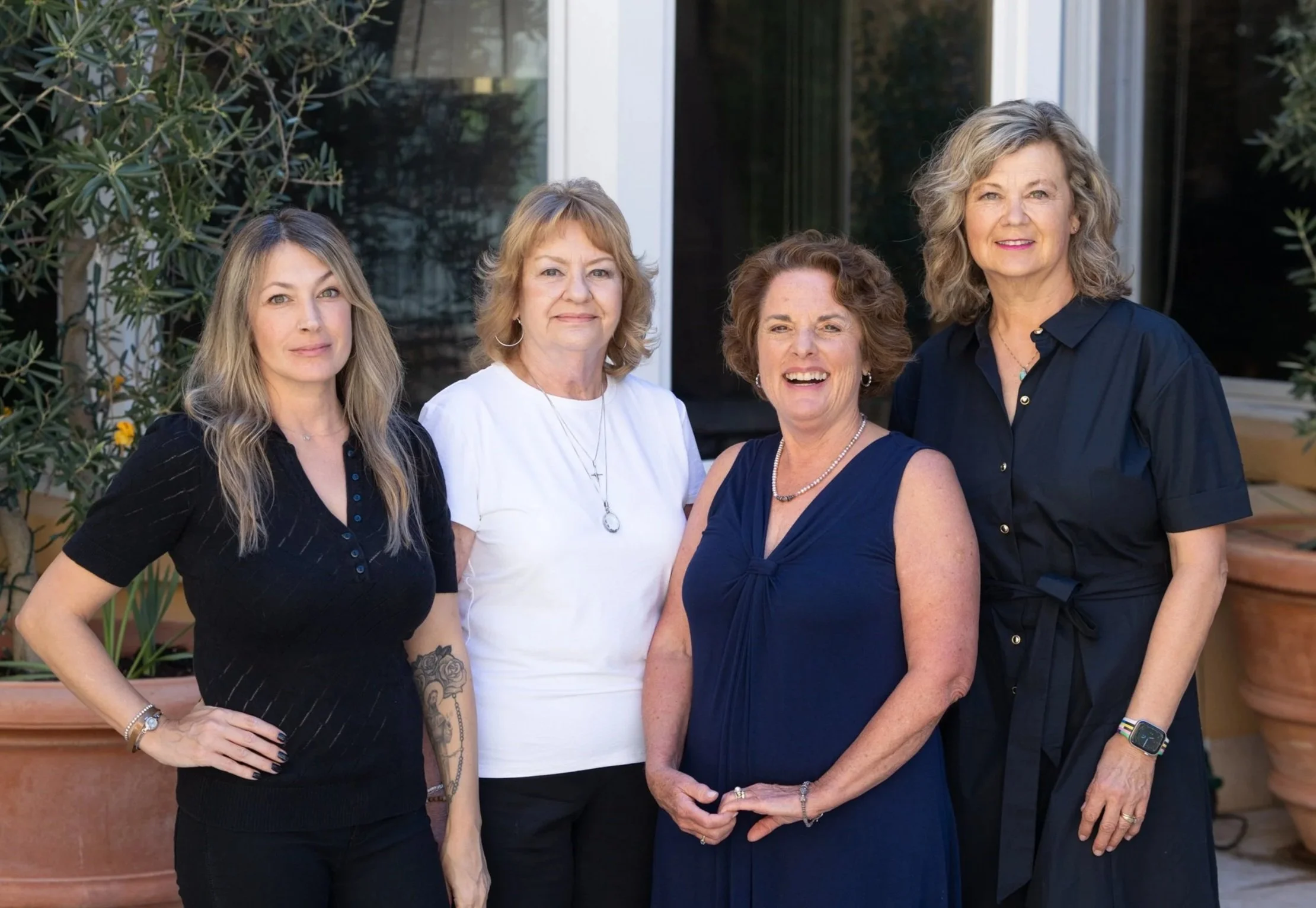 Five women standing side by side outside near a window and greenery, smiling at the camera.