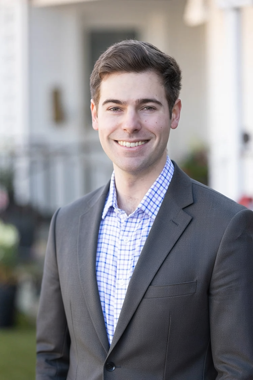 Young man in a gray suit and blue checkered shirt standing outside, smiling, in front of a blurred residential neighborhood background.
