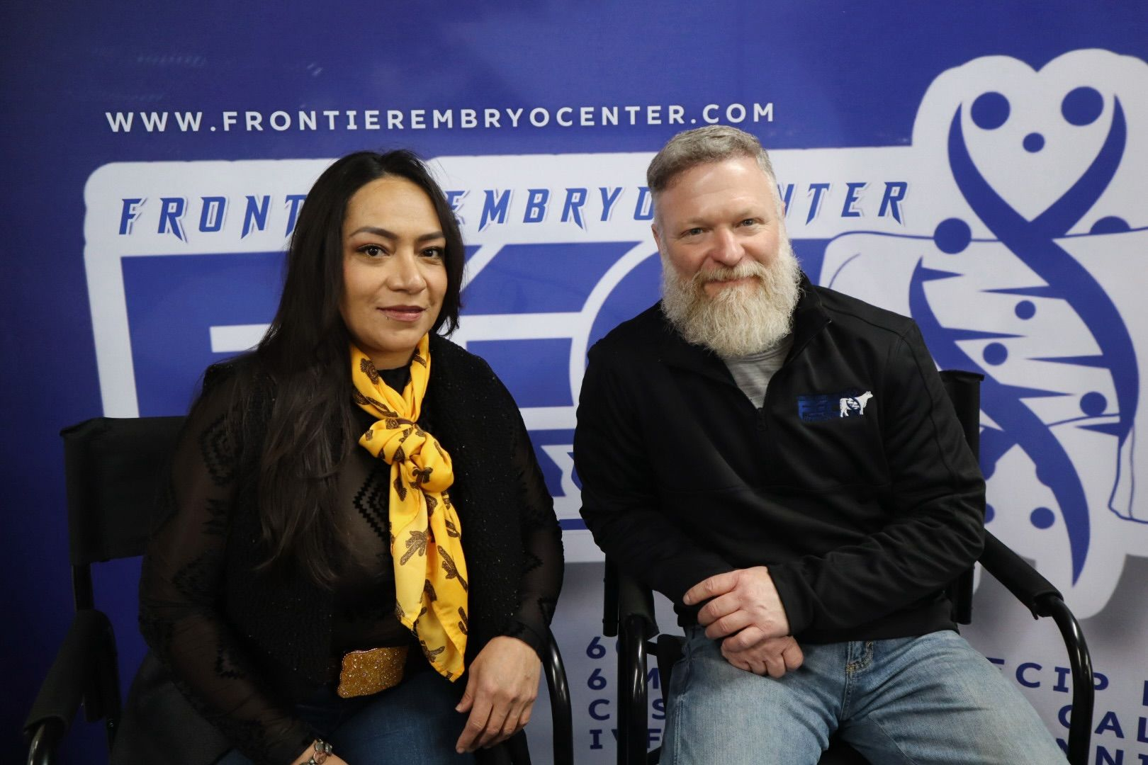 The owners of Caprock Cattle Co standing on a dirt ground in front of large metal grain silos, with a clear sky in the background.