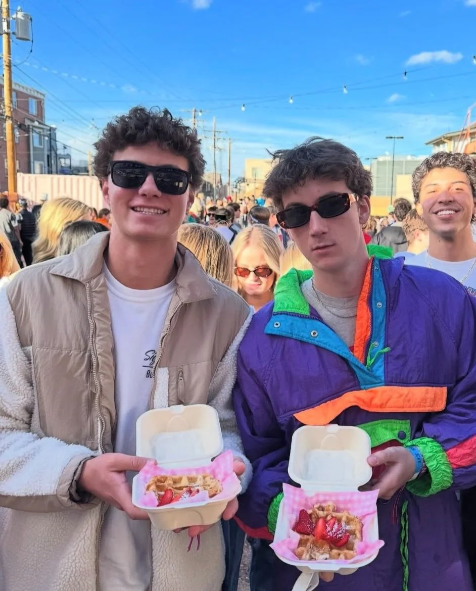 Two people holding takeaway boxes with waffles and strawberries, wearing sunglasses and smiling at an outdoor event.