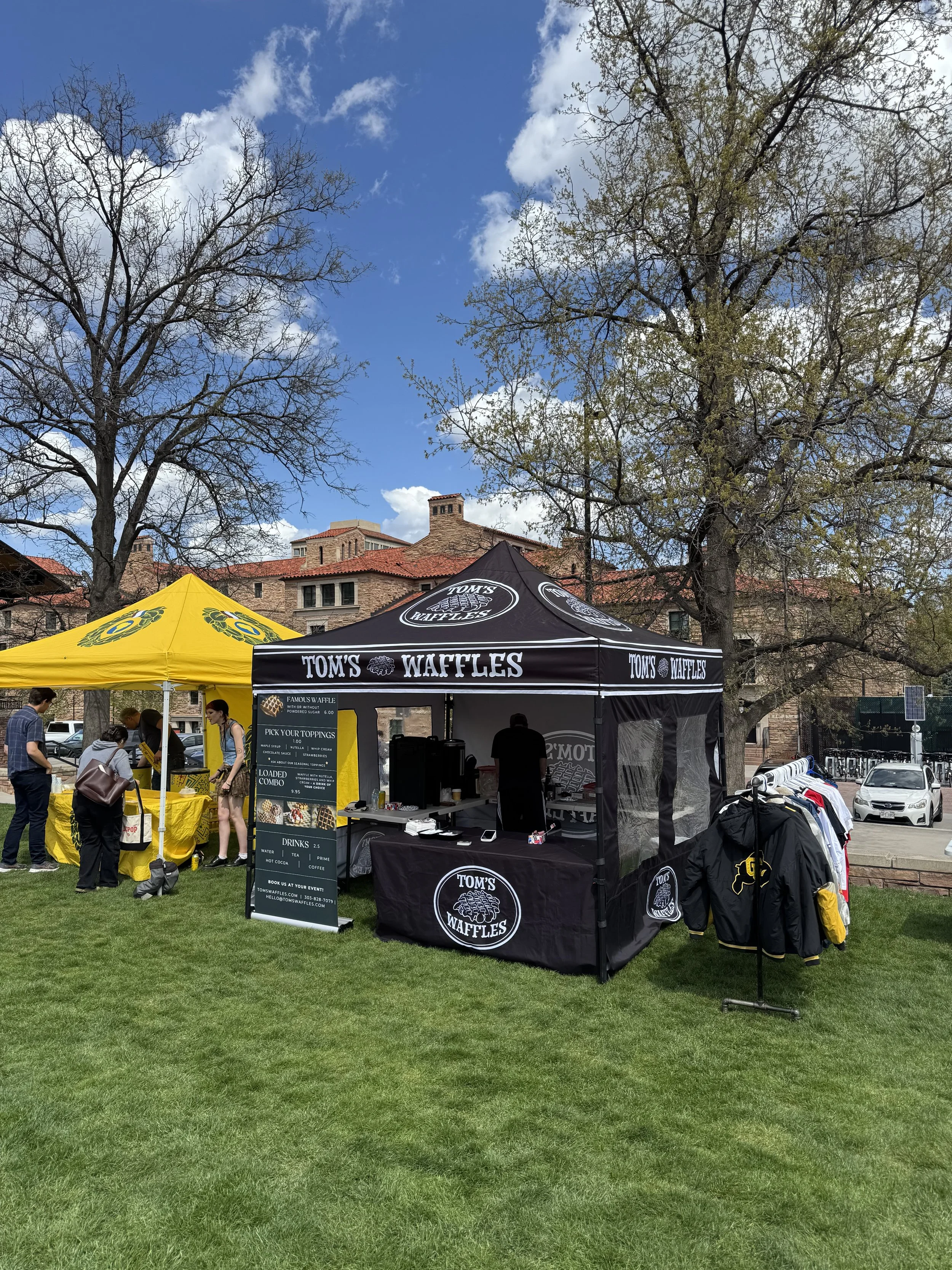 Outdoor event with tents for Tom's Waffles and another vendor, surrounded by trees, grass, and a blue sky.