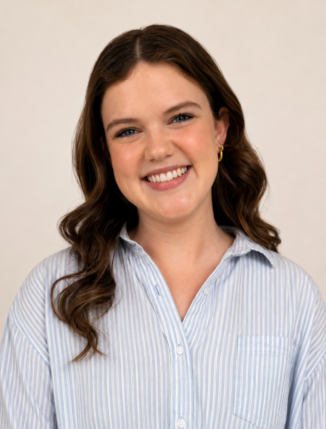 A smiling woman with wavy brown hair and blue eyes, wearing a light blue striped shirt and gold hoop earrings, posed against a plain off-white background.
