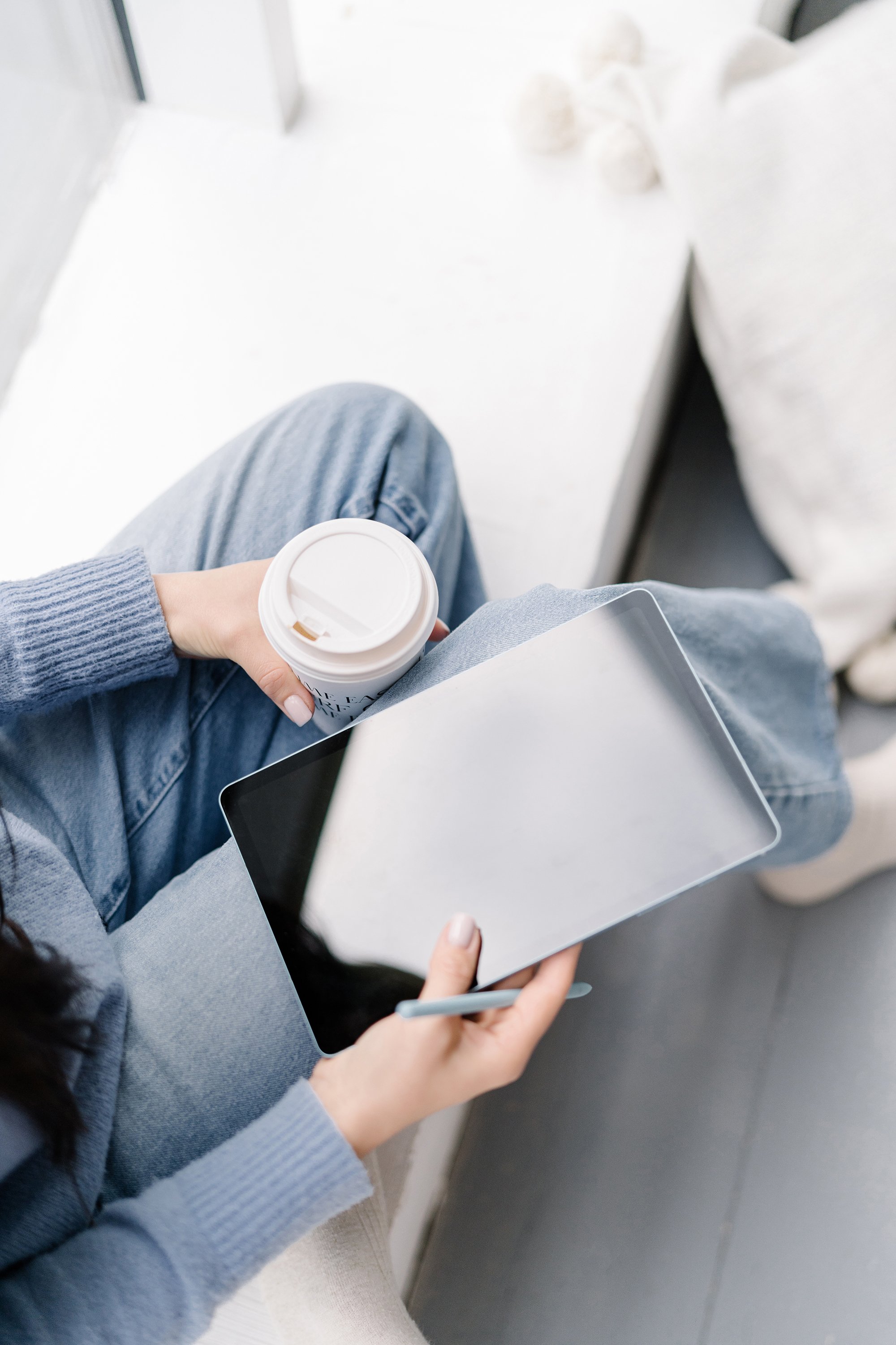 A person sitting on a white bench, holding a disposable coffee cup in one hand and a tablet and stylus in the other. The individual is wearing a blue sweater and jeans, with a white pillow on the bench.