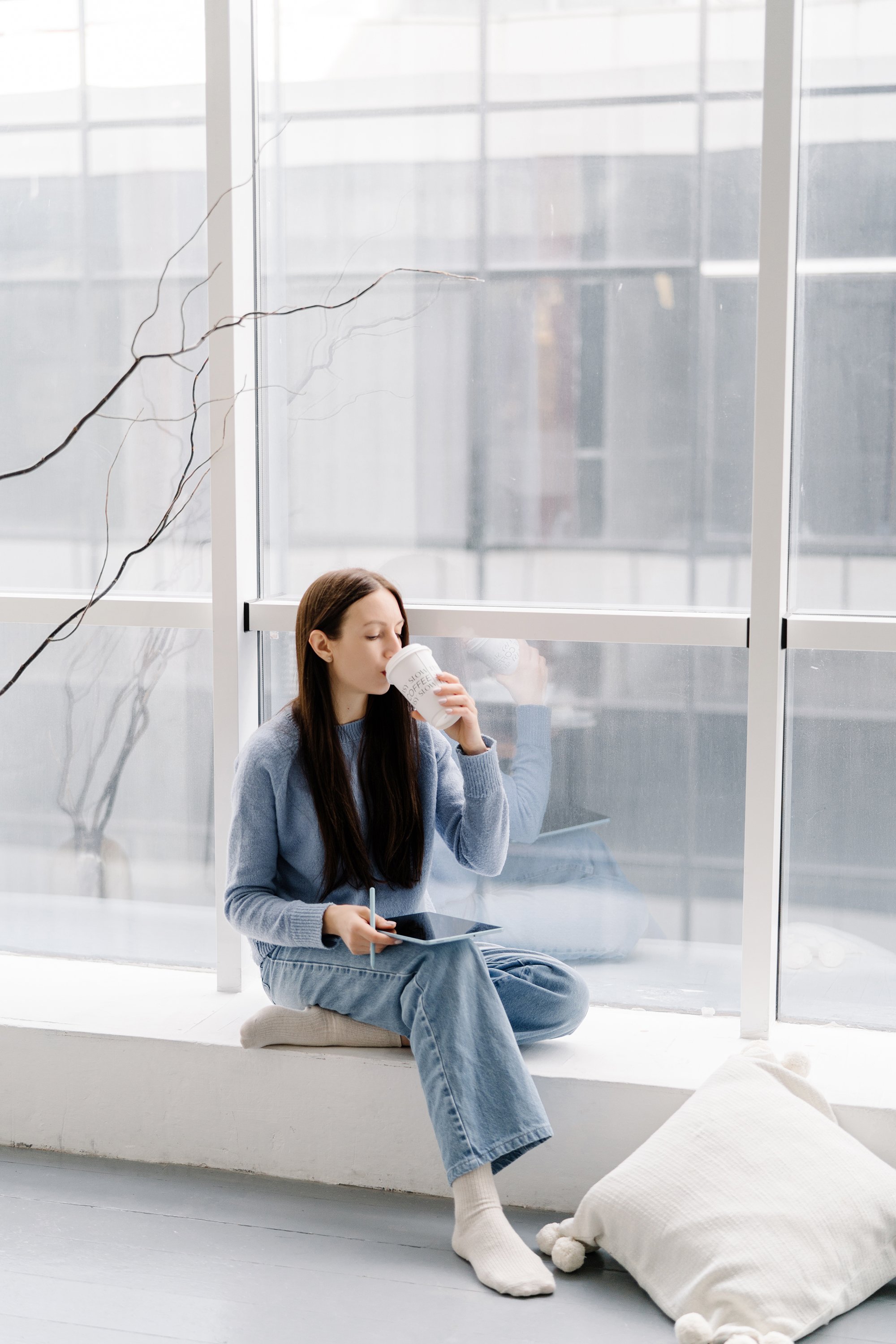 A woman with long brown hair, wearing a gray sweater and blue jeans, sitting on a cushion by a large window. She is drinking coffee from a white cup and holding a tablet with a stylus. There is a white pillow with pom-poms on the floor, and the background shows a cityscape through the window.