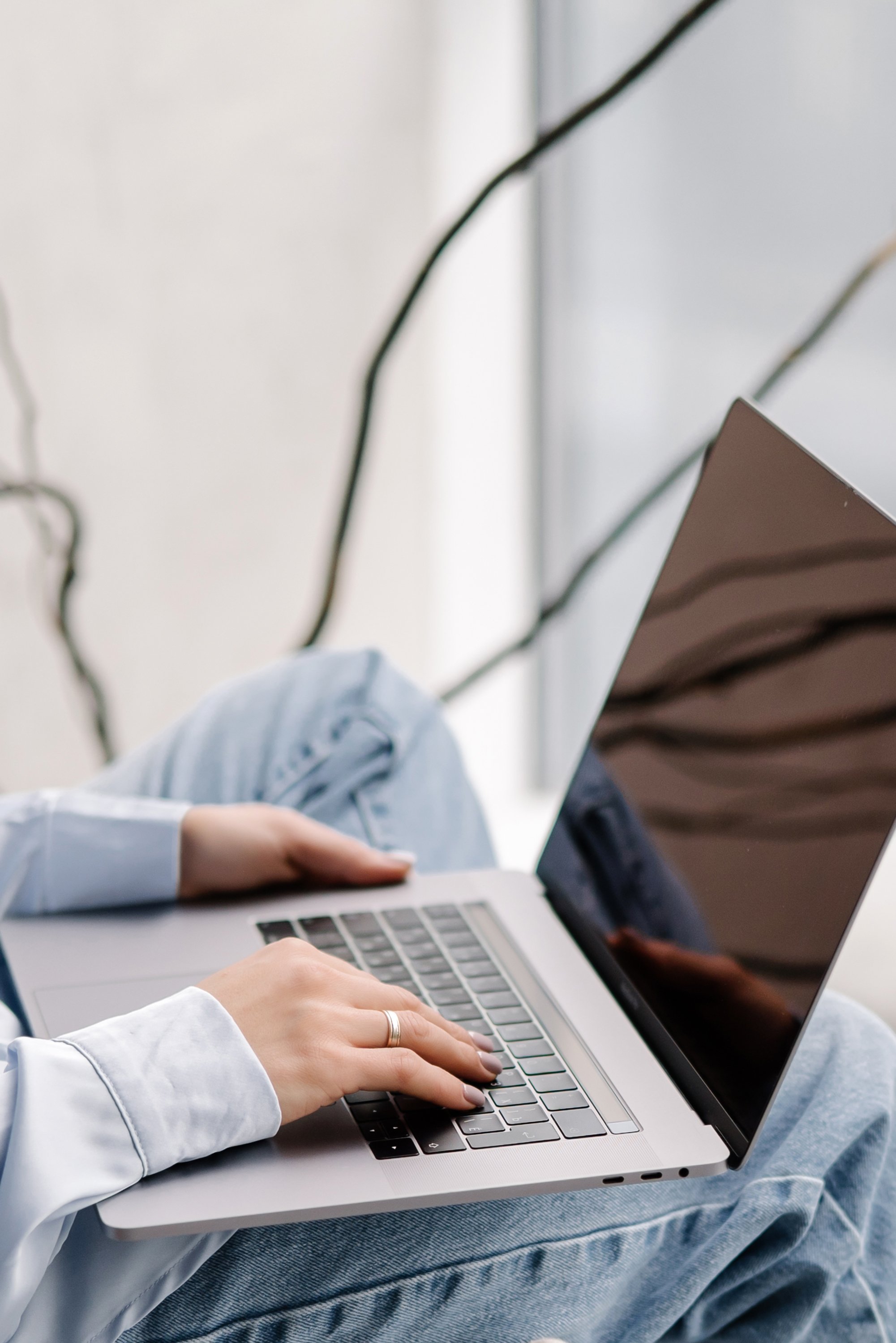 Person wearing a white shirt and blue jeans typing on a silver laptop with a black screen, sitting on a white surface.