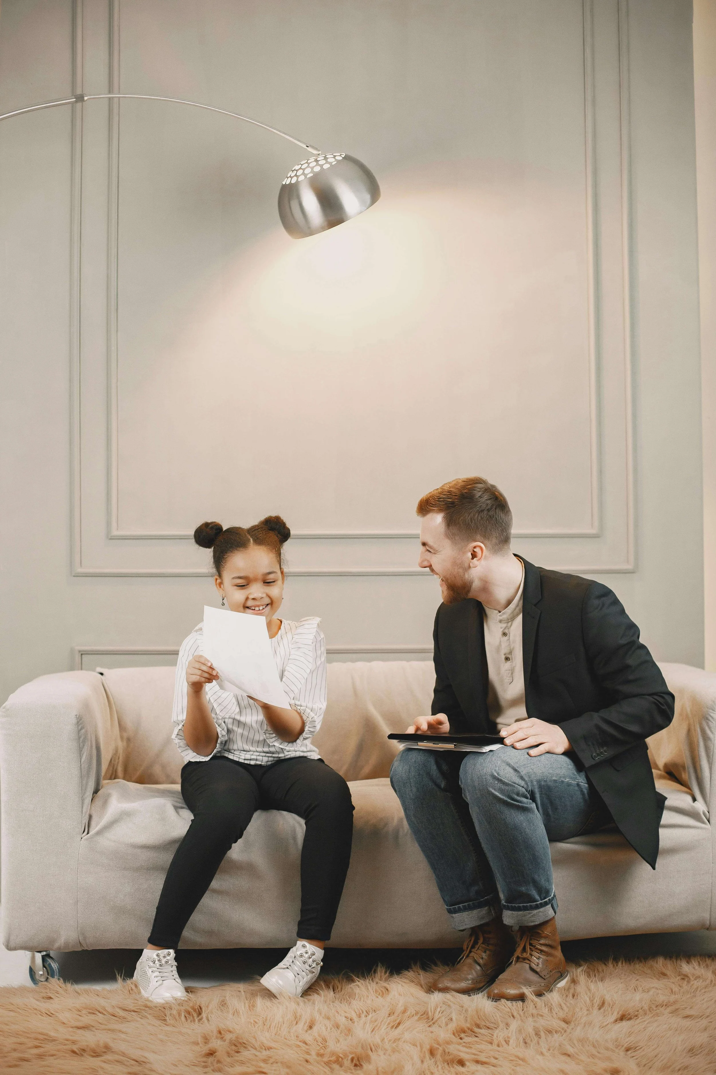 A young girl with a paper in her hands sitting on a beige sofa, smiling, with a man in a black jacket sitting next to her on a couch, holding a tablet, in a modern living room with gray walls and a beige carpet.