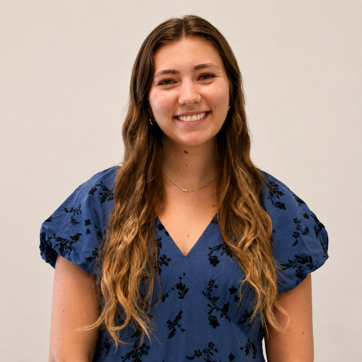 A young woman with long wavy brown hair smiling, wearing a blue floral blouse and gold jewelry, standing against a plain off-white background.