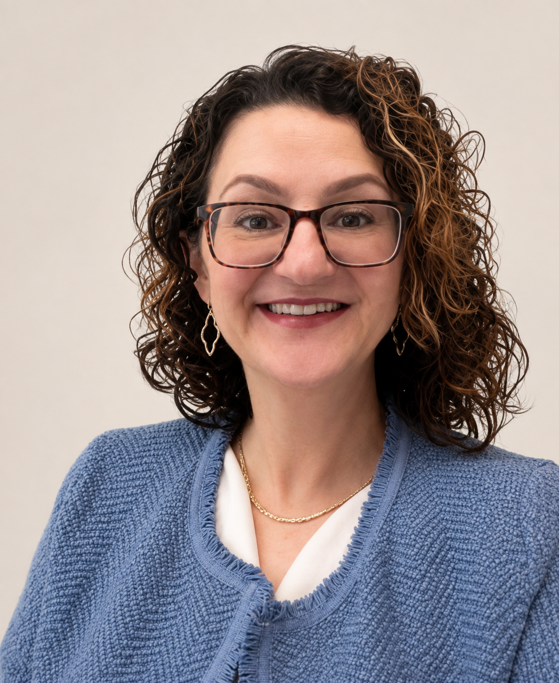 Close-up of a smiling woman with curly brown hair, wearing glasses, gold earrings, a gold necklace, and a blue textured cardigan over a white top against a plain light background.