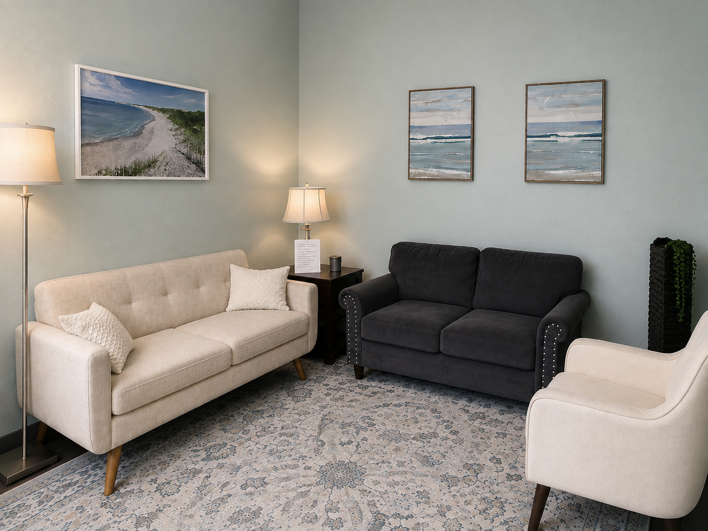 Living room with three sofas, two white and one black, a side table with a lamp, framed ocean-themed artwork, and a patterned rug.