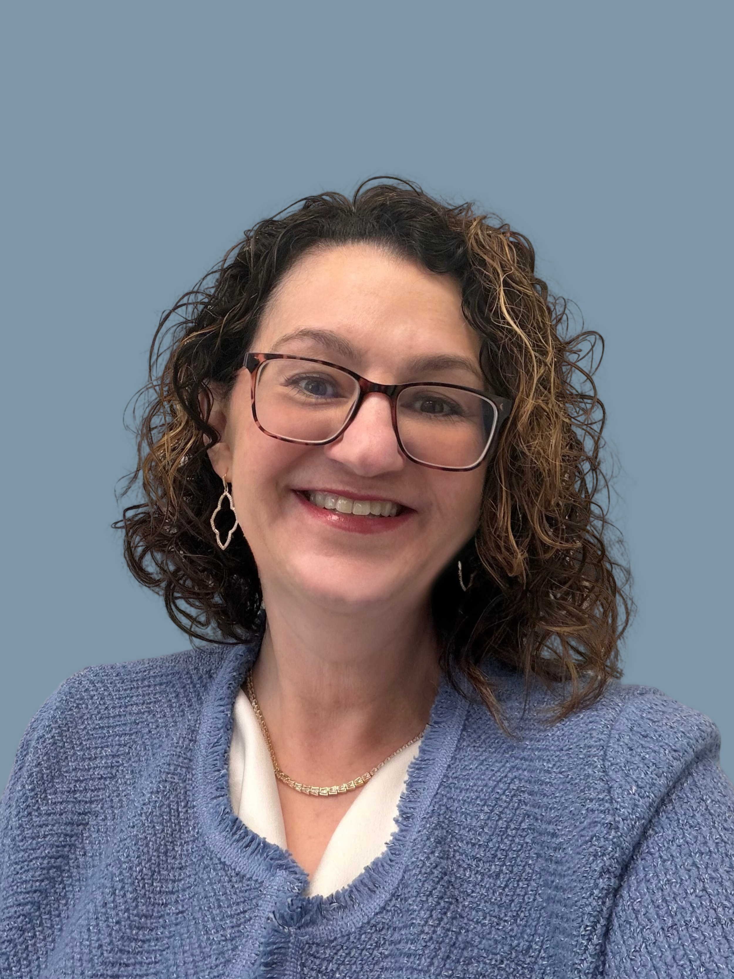 Close-up of a woman with curly brown hair wearing glasses, jewelry, a white top, and a blue sweater, smiling against a solid light blue background.