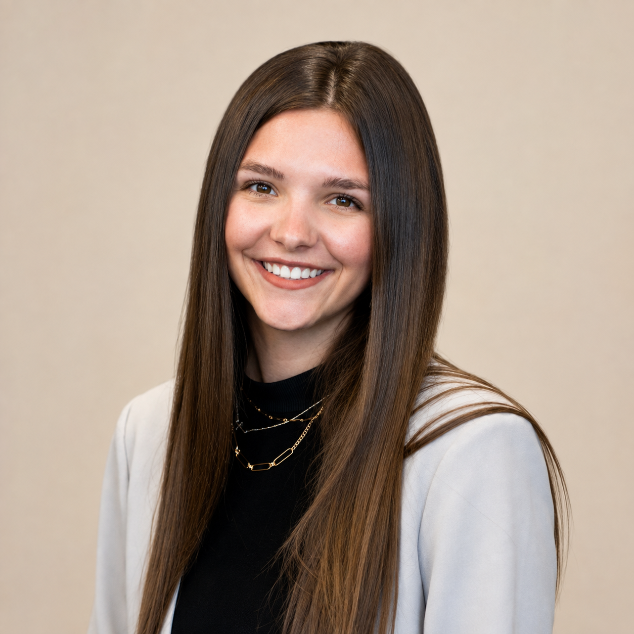 Portrait of a young woman with long brown hair, smiling, wearing a white blazer, black top, and layered gold necklaces, against a plain beige background.