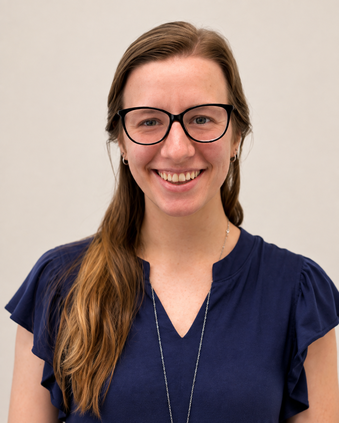 A woman with light skin and long wavy red hair, wearing black glasses and a navy blue blouse, smiling at the camera against a plain light background.