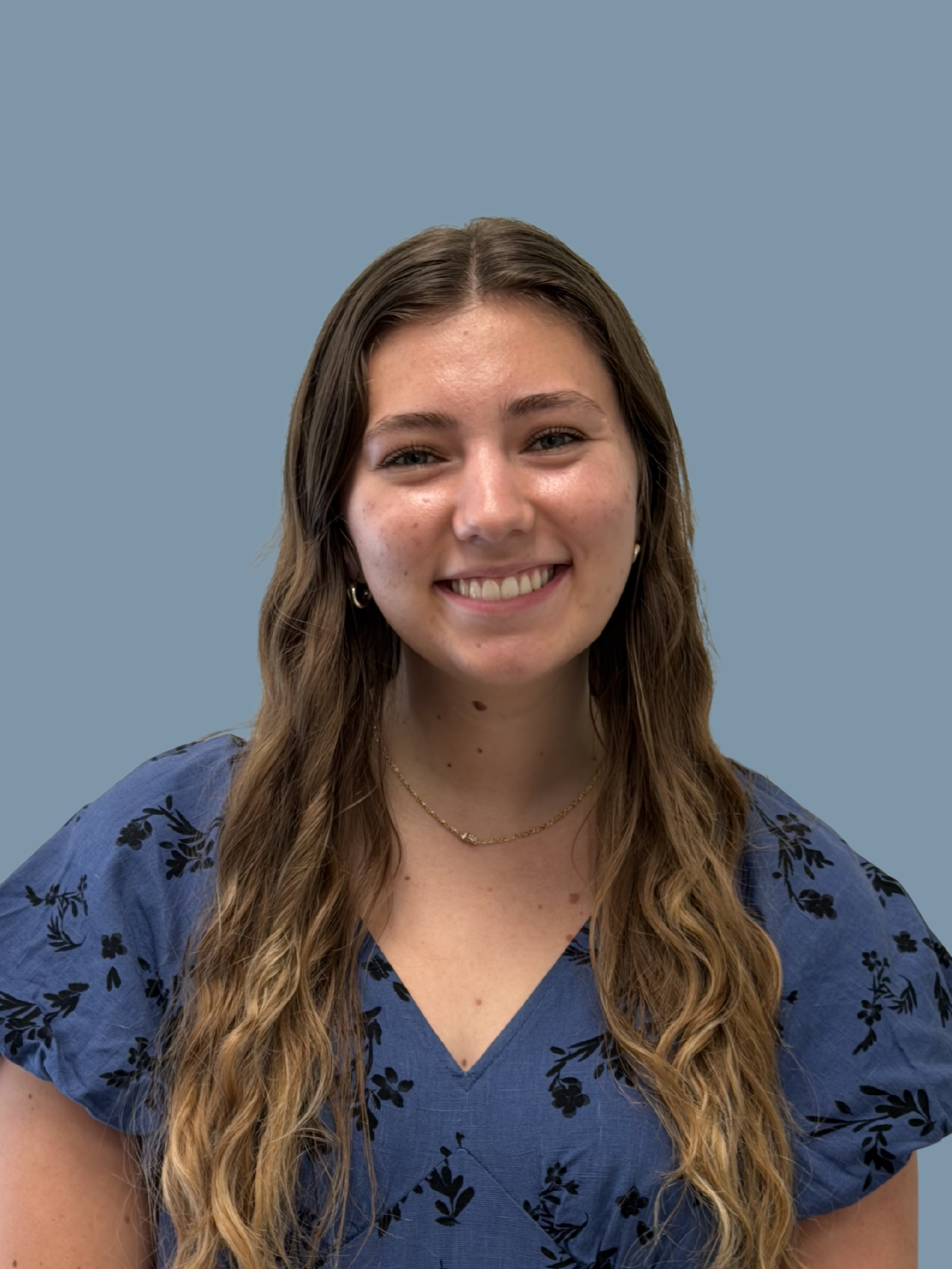 Close-up of a smiling young woman with long, wavy brown hair, wearing a blue floral dress, against a light blue background.