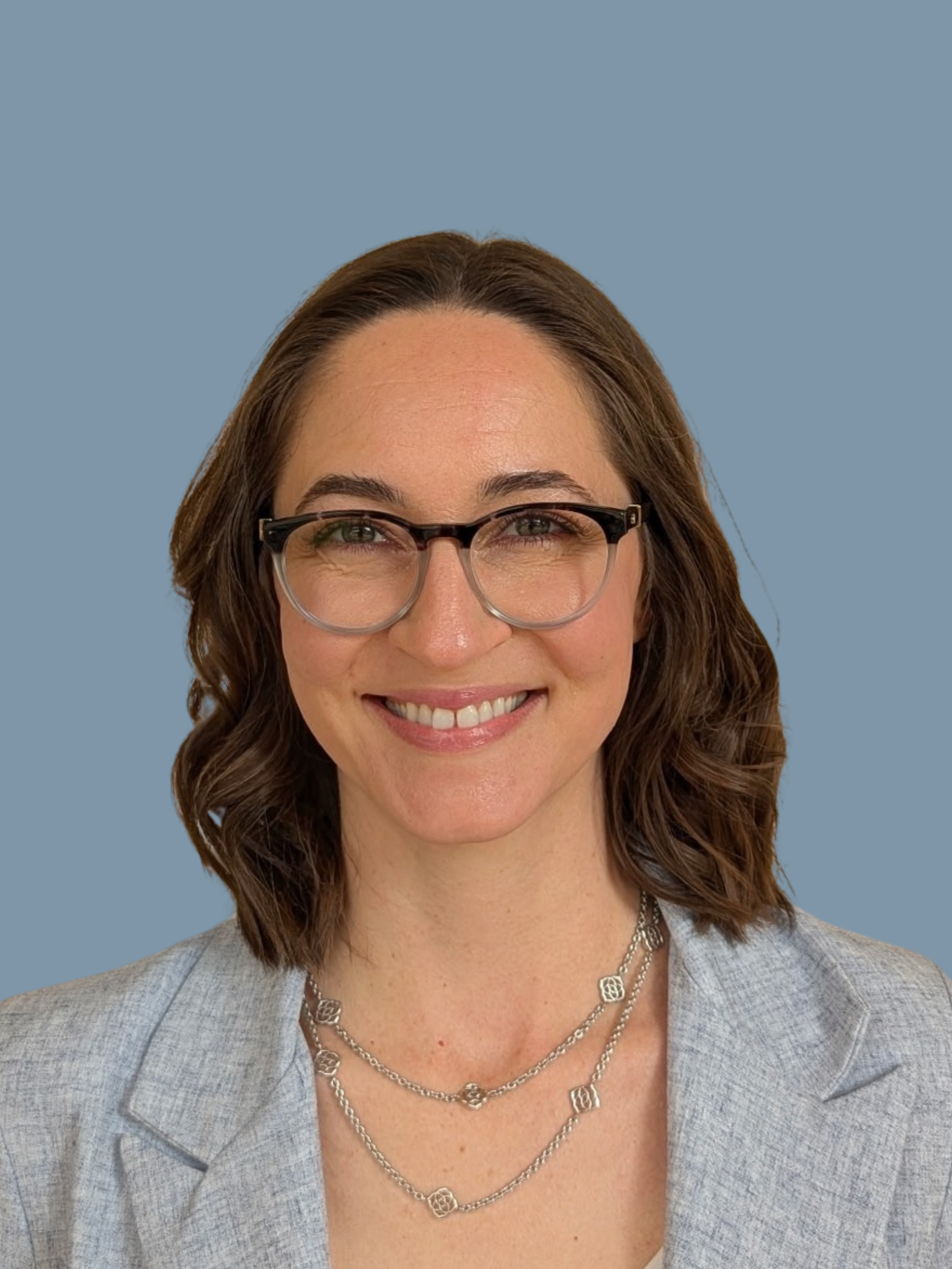 Close-up of a woman with brown hair, glasses, smiling, wearing a gray blazer and layered silver necklaces, against a plain blue background.