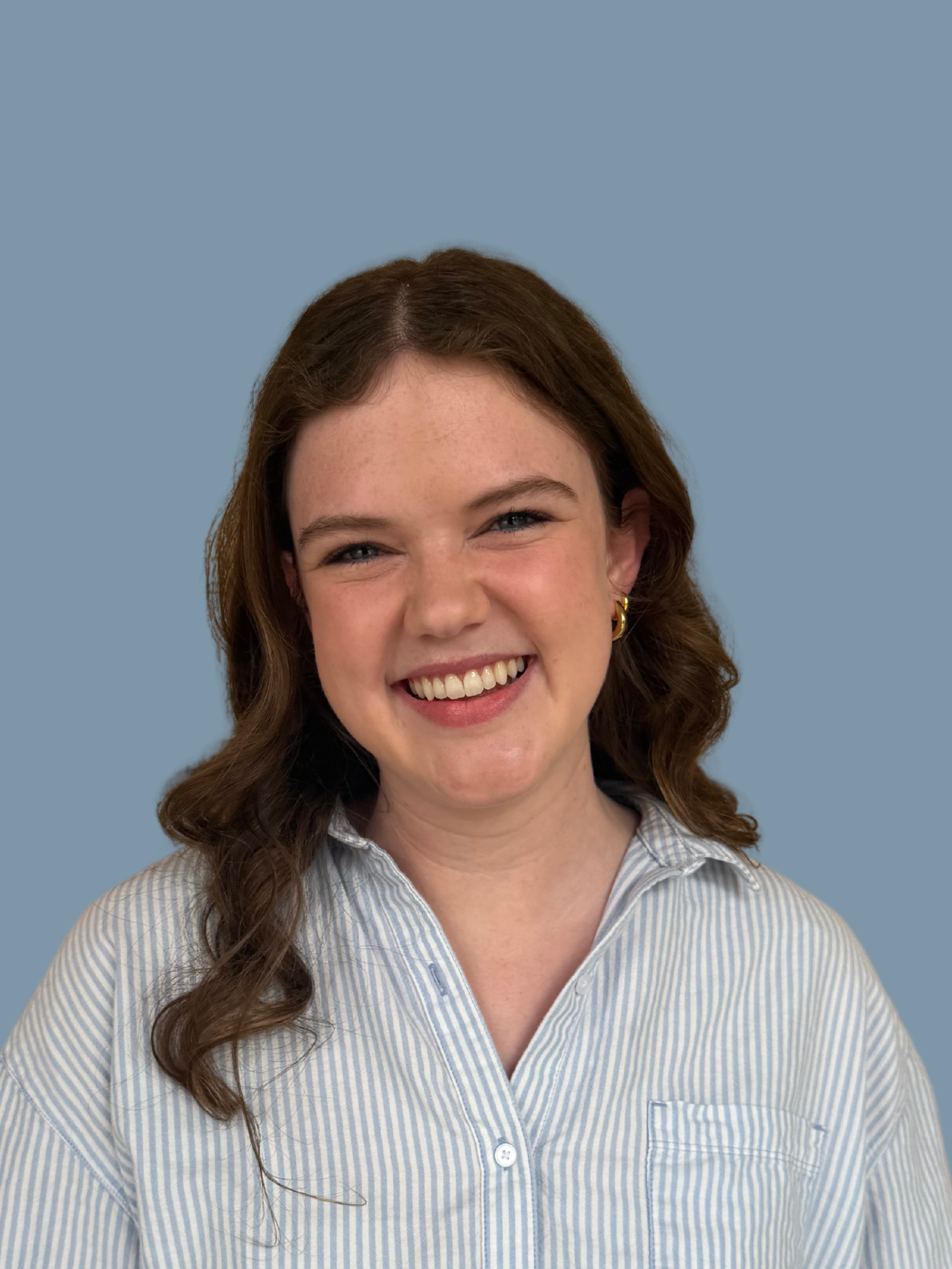 Close-up of a smiling woman with long wavy brown hair wearing a light blue and white striped shirt against a plain light blue background.