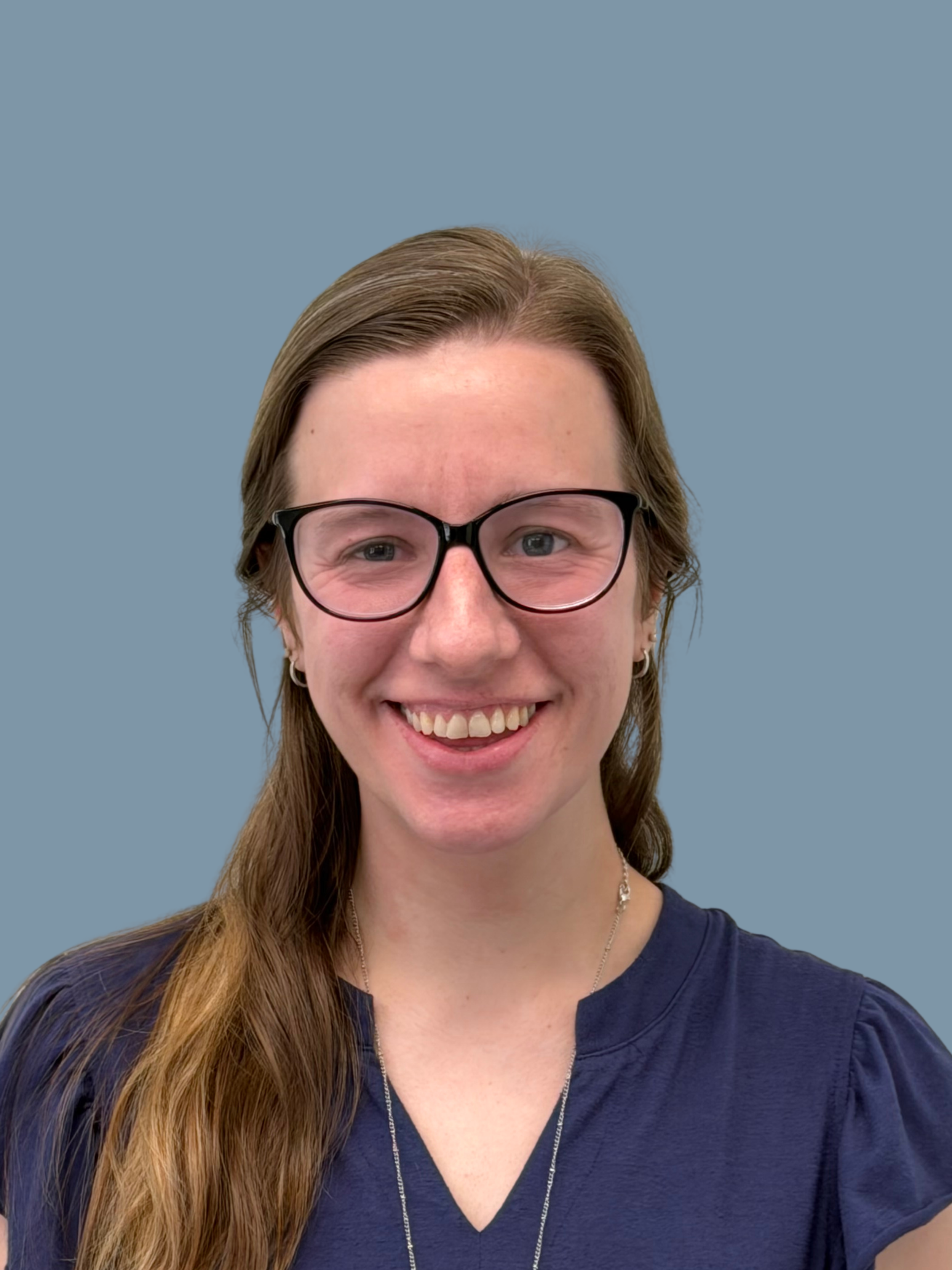Close-up of a smiling woman with long, brown hair, glasses, earrings, wearing a navy blue top and a necklace, against a plain light blue background.