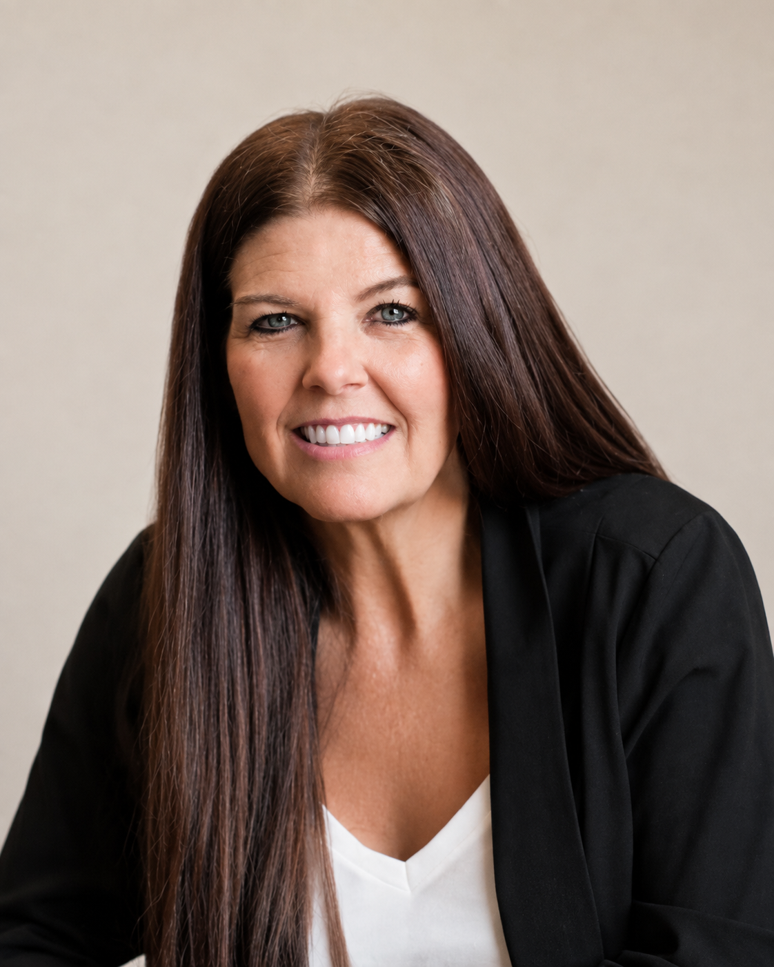 A woman with long brown hair, blue eyes, and a bright smile, wearing a white top and a black blazer, posed against a plain beige background.