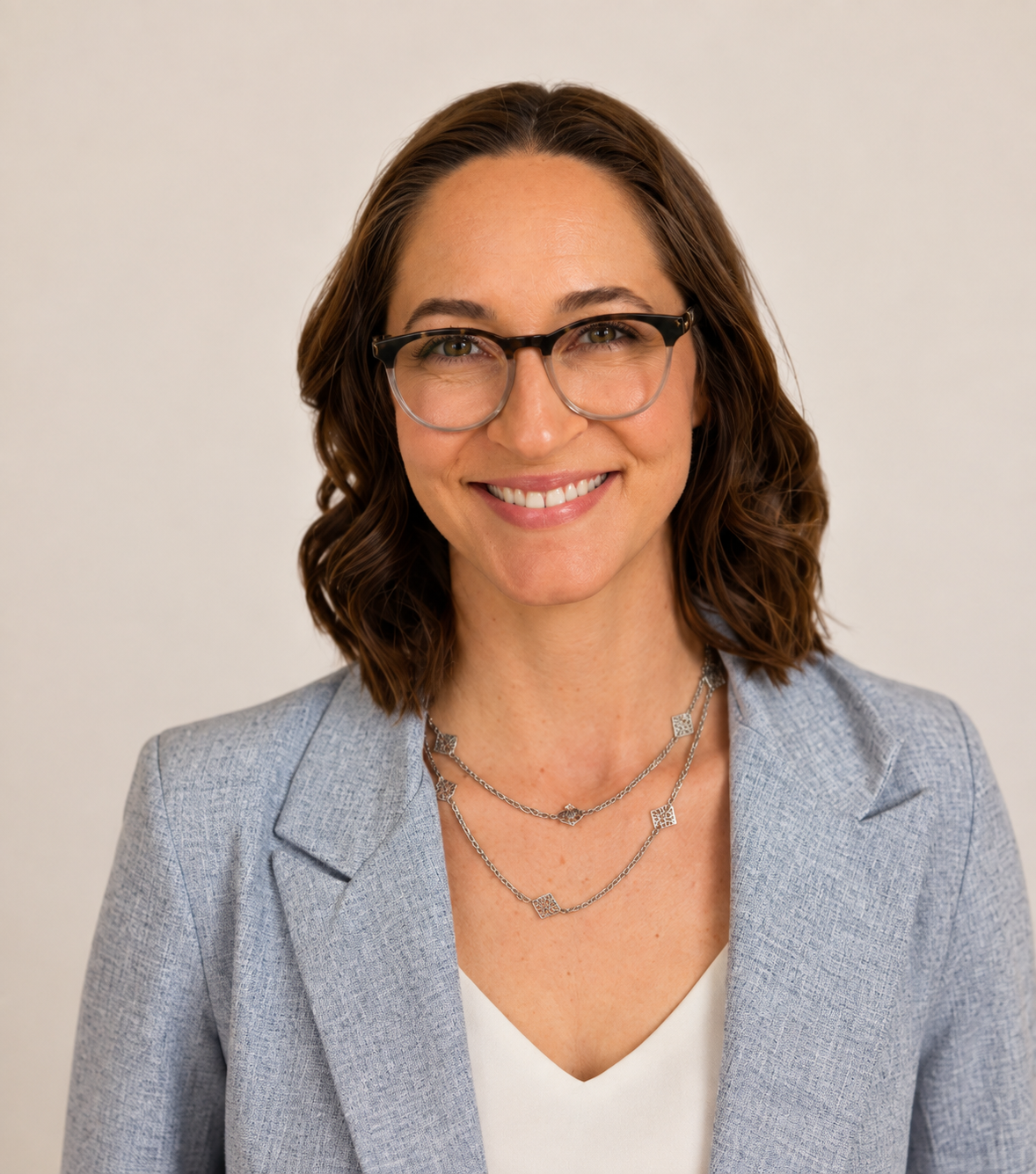 A woman with shoulder-length wavy brown hair, glasses, and light skin, wearing a light blue blazer over a white top, smiling against a plain light-colored background.