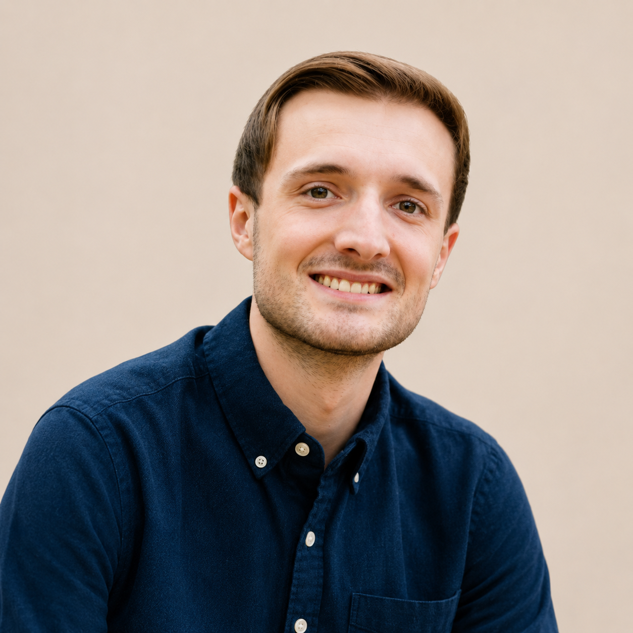 Portrait of a young man with light brown hair, smiling, wearing a navy blue button-up shirt, against a plain beige background.