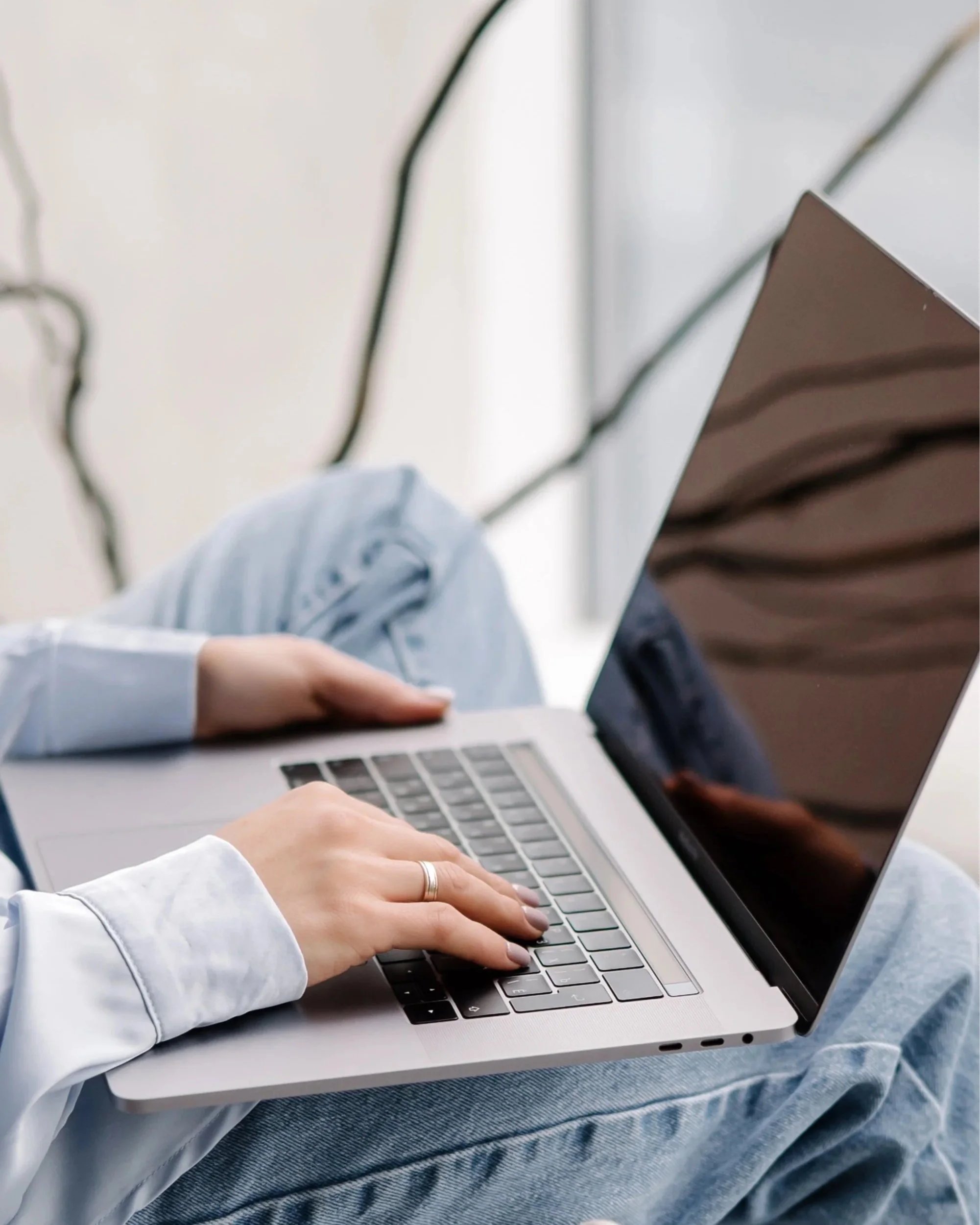 A person wearing a light-colored shirt and jeans sitting cross-legged on a surface, using a silver laptop. The person's hands are on the keyboard, and the laptop screen is open, reflecting part of their face and surroundings.