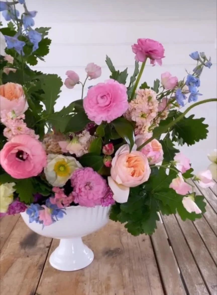 A white ceramic vase filled with pink, peach, and blue flowers on a wooden table.