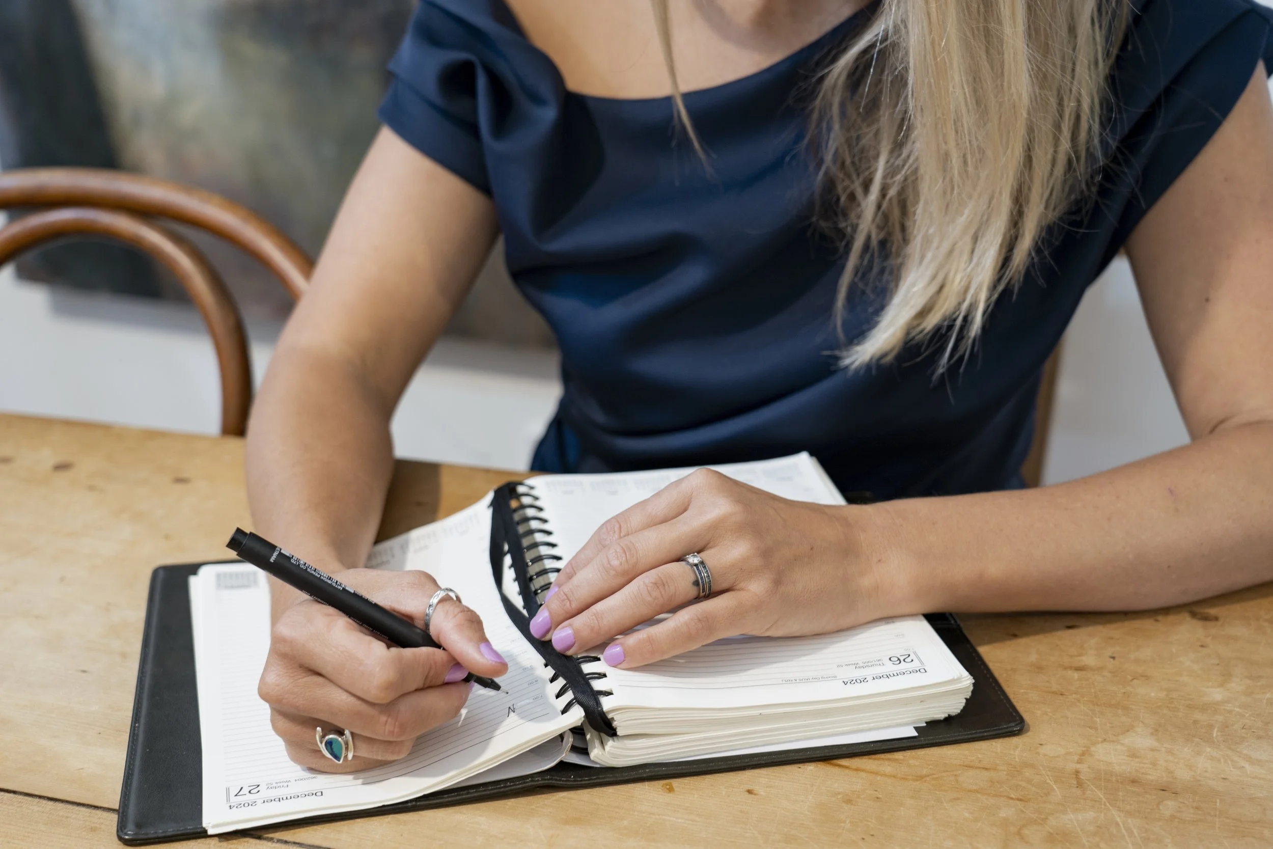 Woman with long blonde hair wearing a navy blue top, writing in a planner on a wooden table.