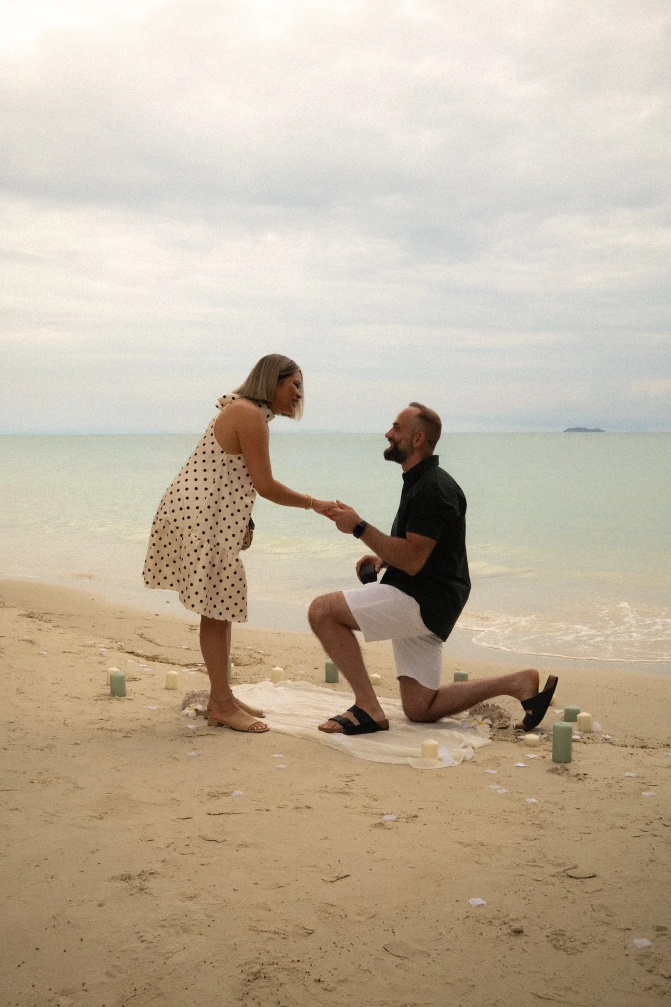 A man proposing marriage to a woman on the beach during sunset, with a large ocean and a distant island in the background. The man is kneeling on one knee, holding the woman's hands, while the woman is standing facing him. There are candles and a whi