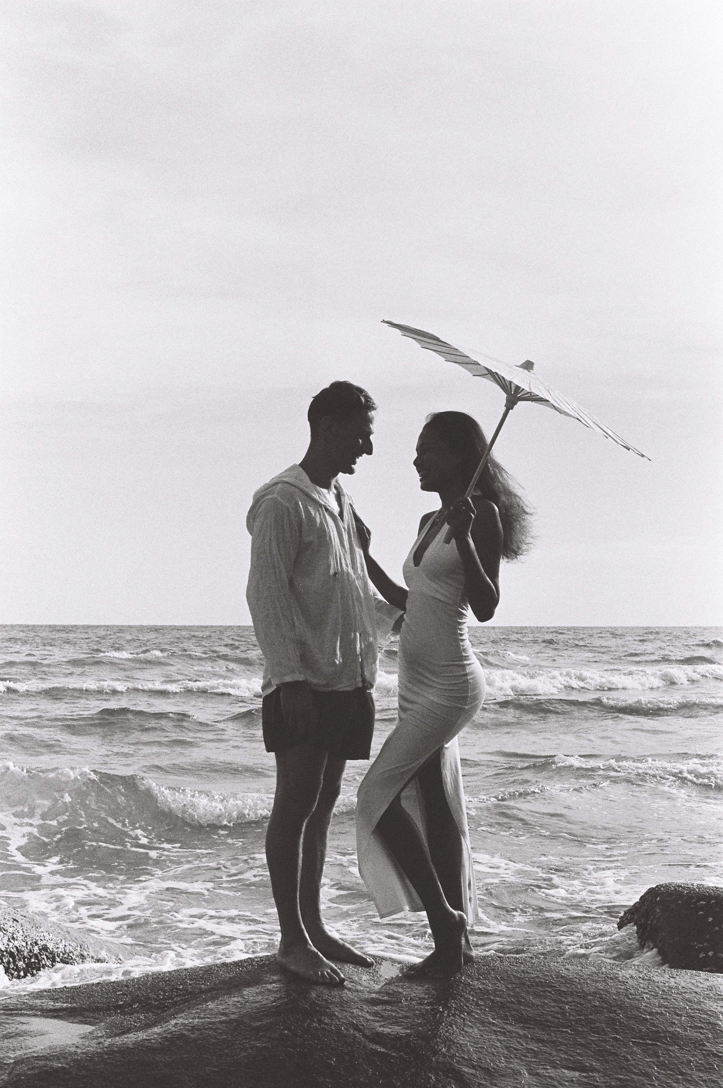 A couple stands on the beach at sunset, holding hands and facing each other. The woman holds an umbrella, and the ocean waves are behind them.