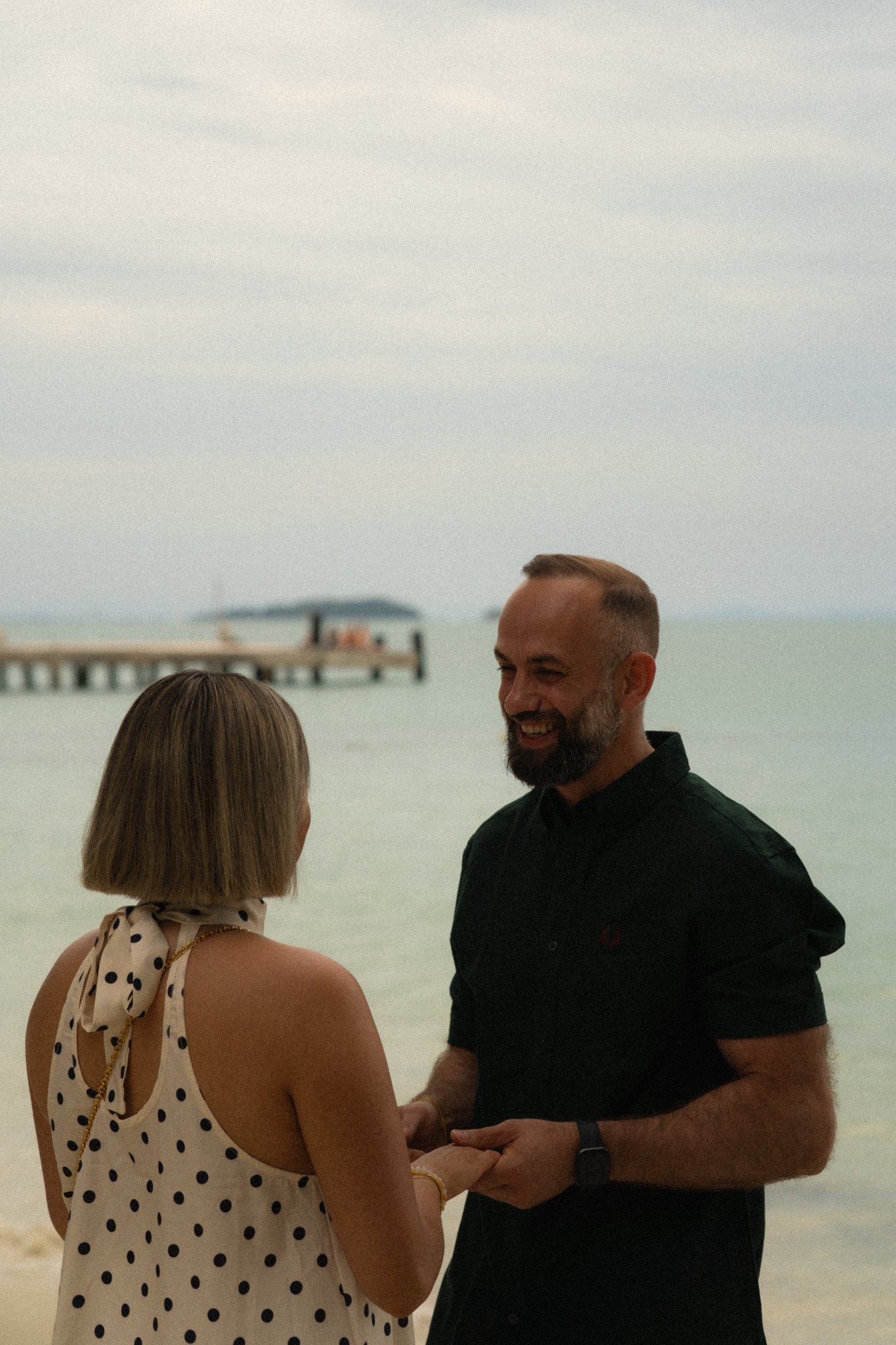 A man and woman are holding hands and facing each other on the beach with a pier in the background, engaging in a conversation. The woman is wearing a sleeveless polka dot dress, and the man is dressed in a black shirt, smiling.