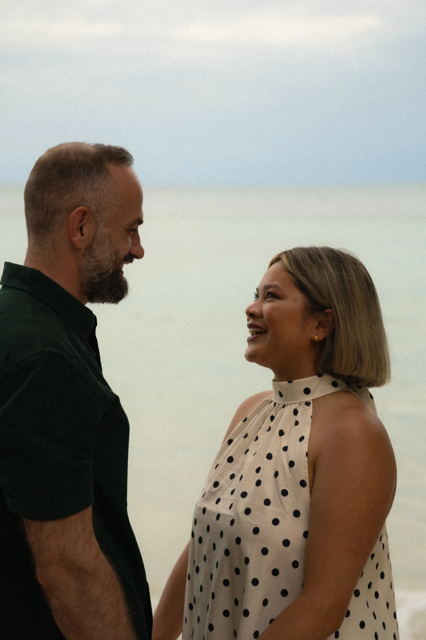 A man and woman are facing each other smiling, holding hands, with a calm ocean and sky in the background.