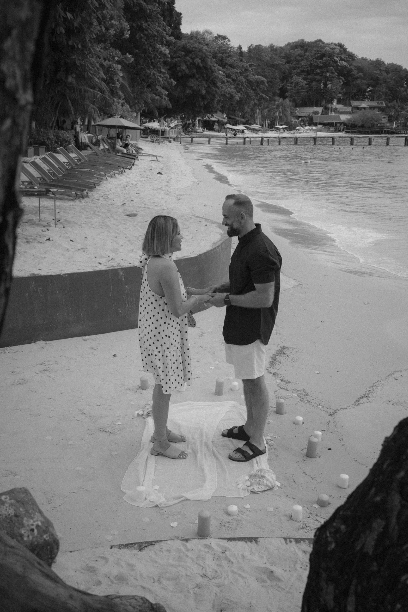 A couple is standing on a beach, holding hands and facing each other. They are in a small, makeshift wedding ceremony with candles on the sand and a white cloth beneath their feet. Behind them, the beach extends along the shoreline with lounge chairs