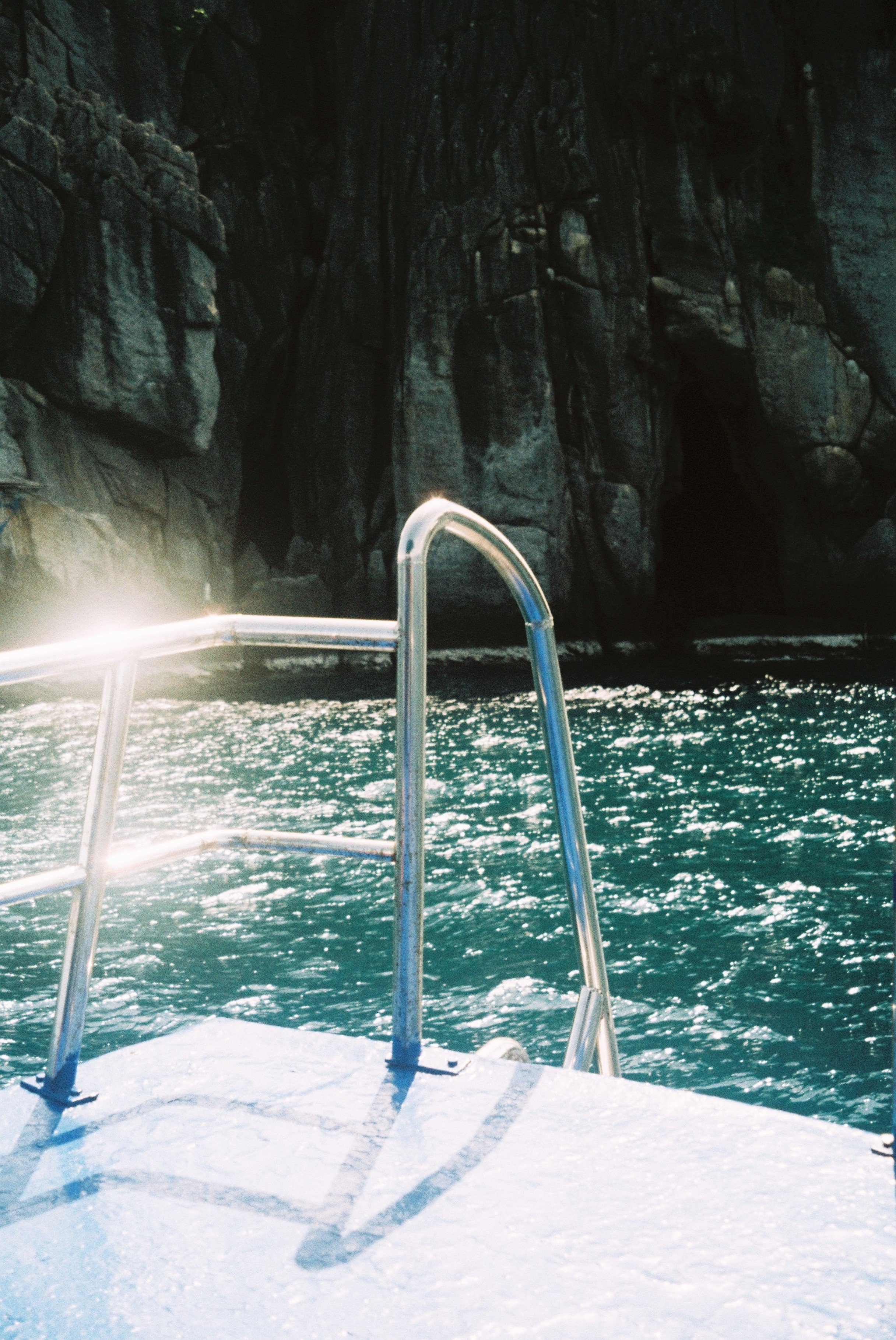 View from a boat showing rocky, dark cliffs and the water below, with a metal railing in the foreground.