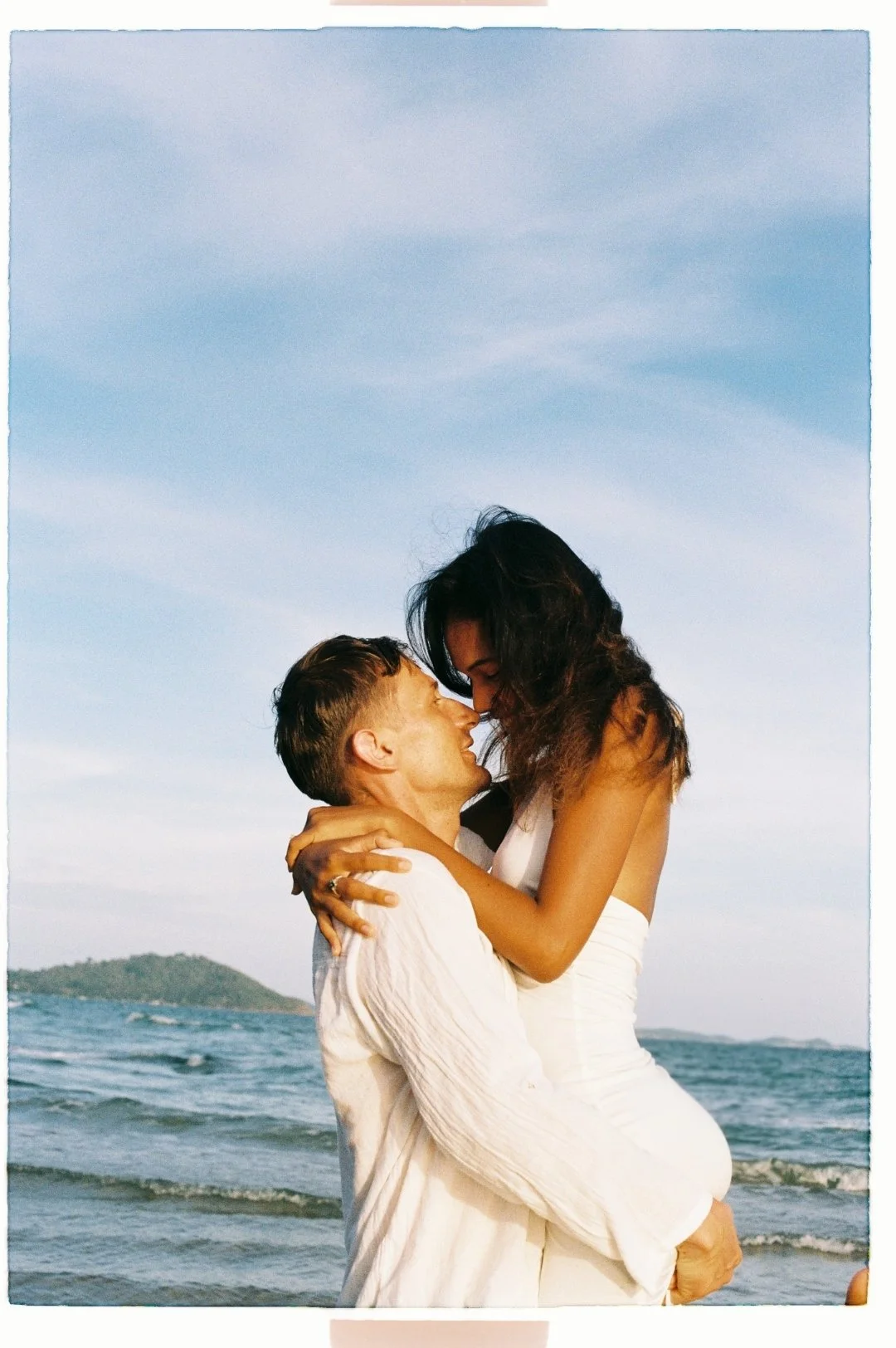 A man is lifting a woman at the beach, and they are touching foreheads while smiling. The background shows the ocean and a distant island under a partly cloudy sky.
